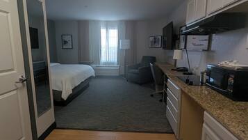 Quarto de hotel com cama, escrivaninha, cadeira, micro-ondas e cozinha compacta.