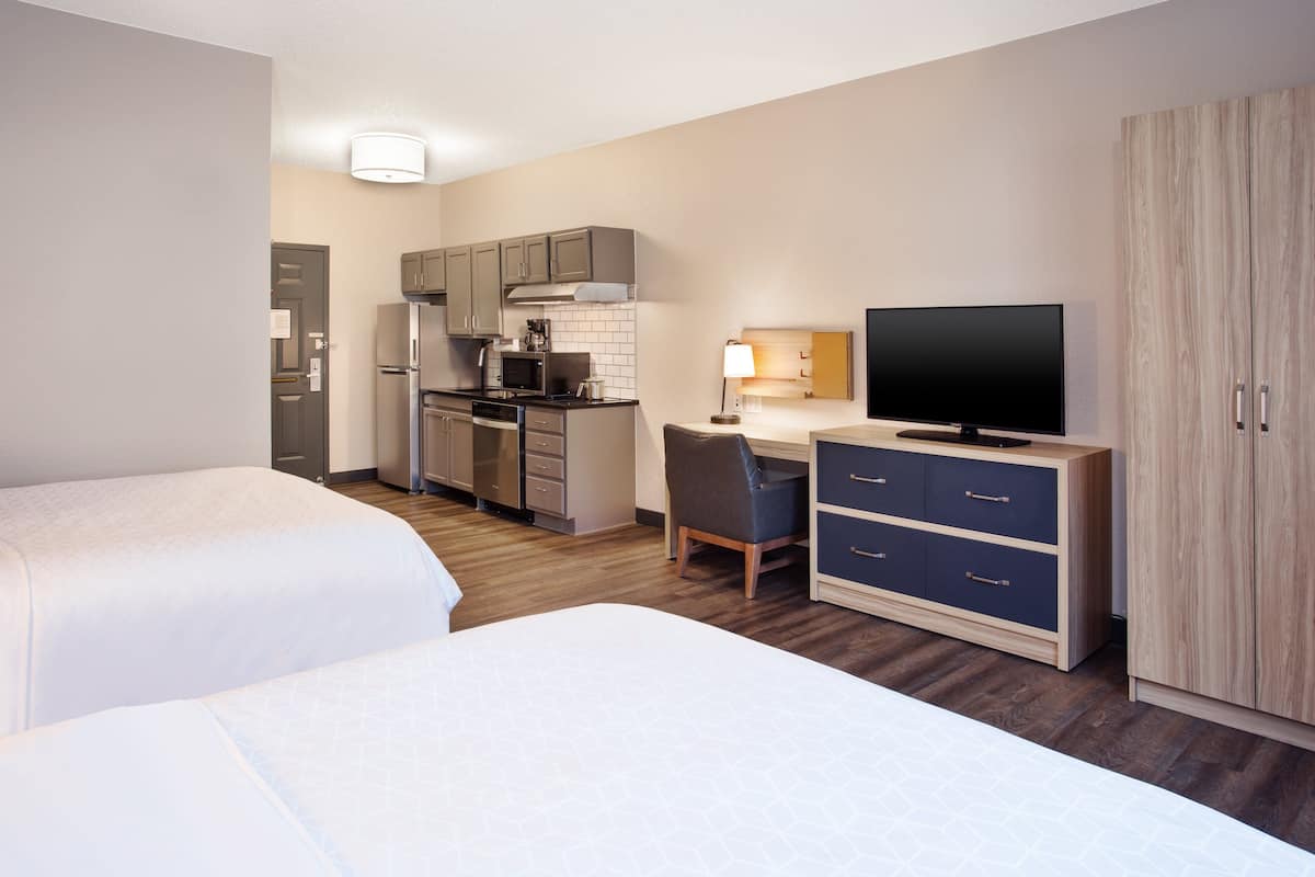 Ein Hotelzimmer mit zwei Betten, einer Küchenzeile, einem Schreibtisch mit Stuhl, einem Fernseher und einem Kleiderschrank.