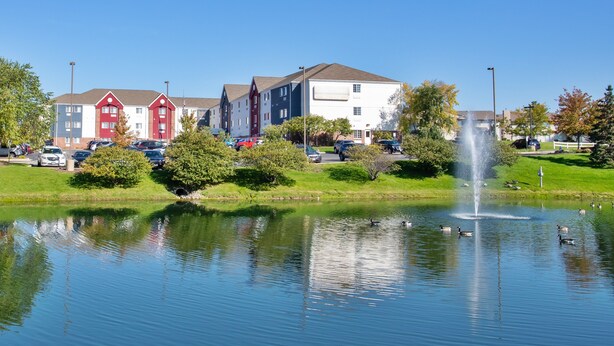 A tranquil pond with a fountain, surrounded by lush greenery and a row of colorful apartment buildings. - Candlewood Suites Kenosha – Pleasant Prairie by IHG (Kenosha)