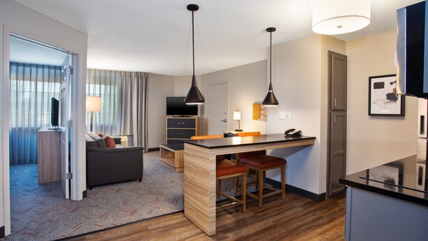 A modern hotel room with a wooden floor, a sofa, a desk with two chairs, a television, and a kitchen area with a refrigerator and a microwave.