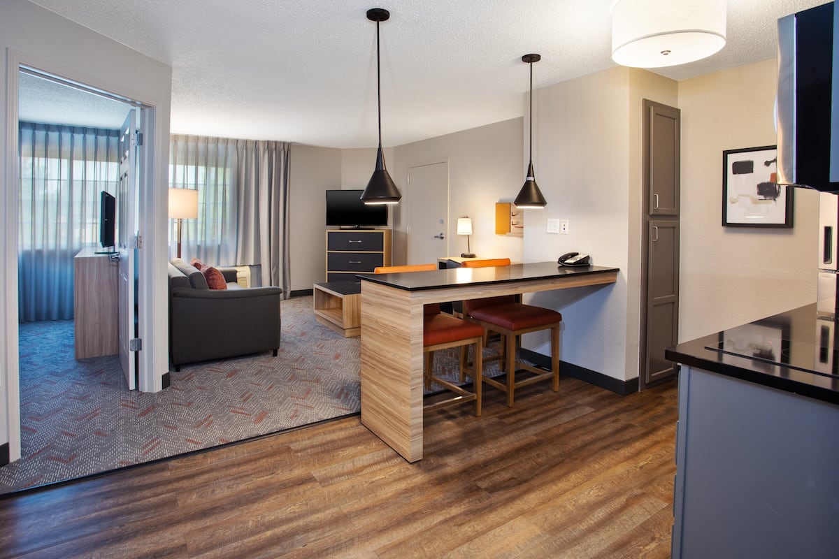 Ein modernes Hotelzimmer mit Holzboden, einer Couch, einem Schreibtisch mit zwei Stühlen, einem Fernseher und einer Küchenzeile mit Kühlschrank und Mikrowelle.
