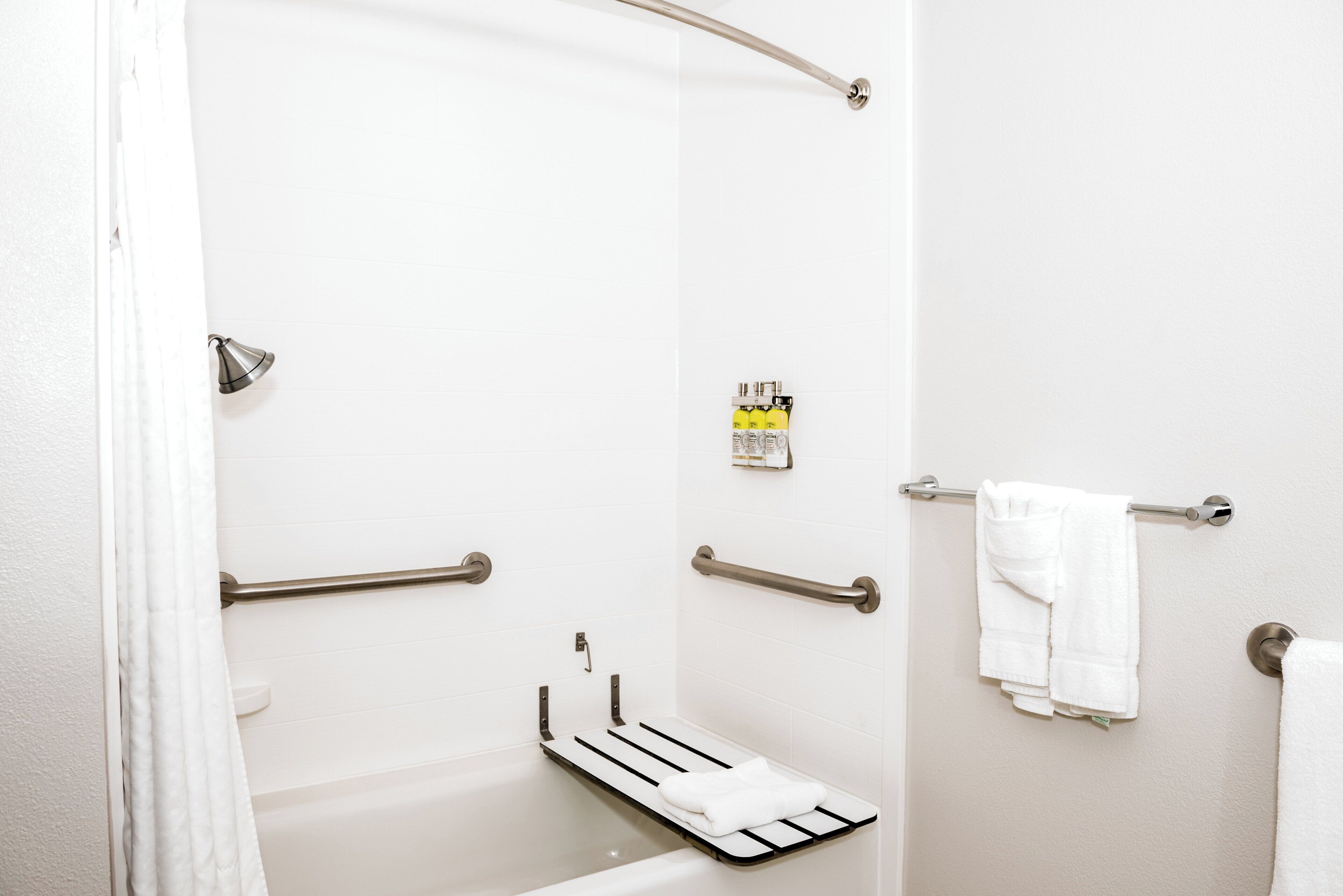 A bathroom with a shower, grab bars, a towel rack, and a soap dispenser.