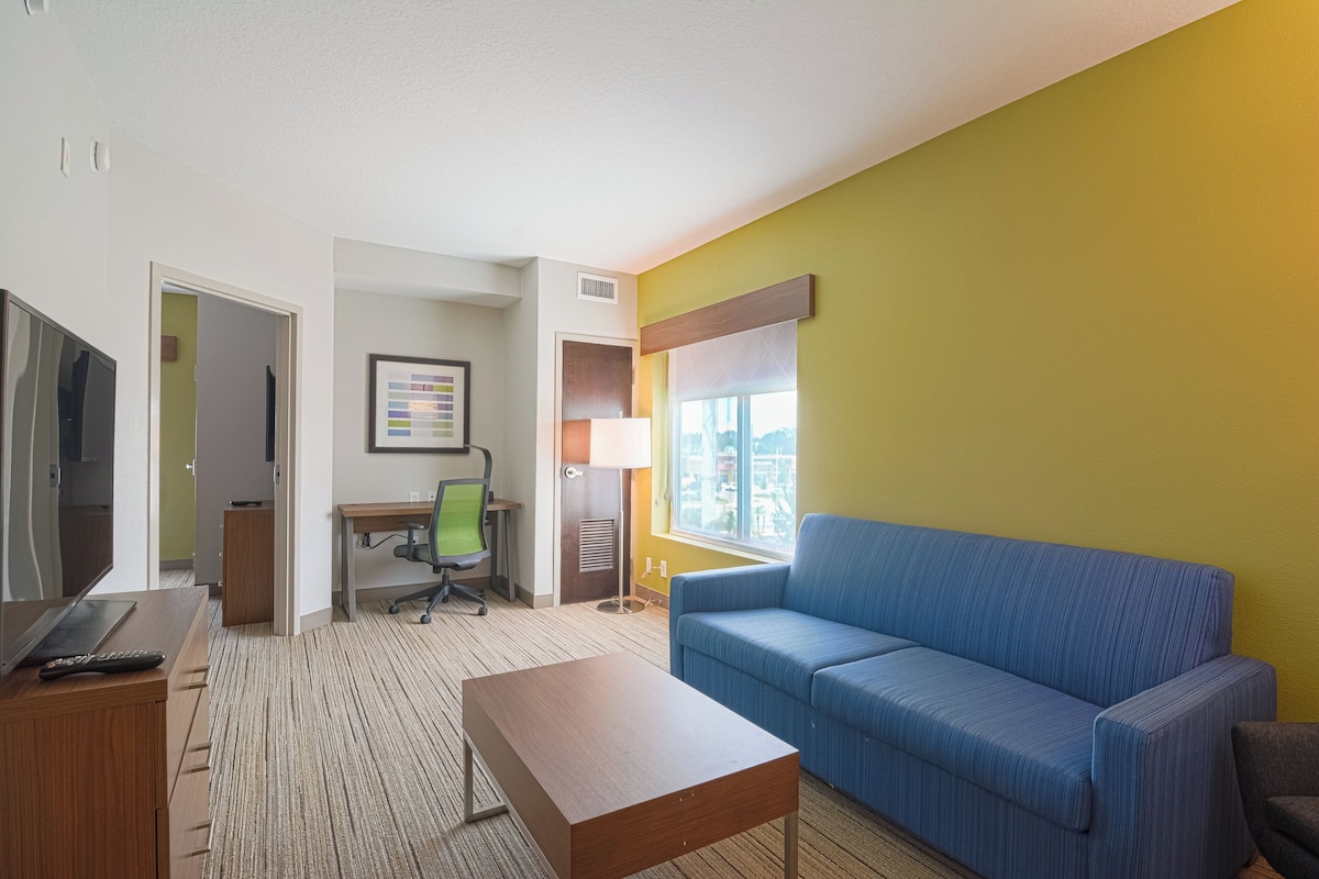 A hotel room with a blue sofa, a wooden desk, a chair, a TV, and a lamp.