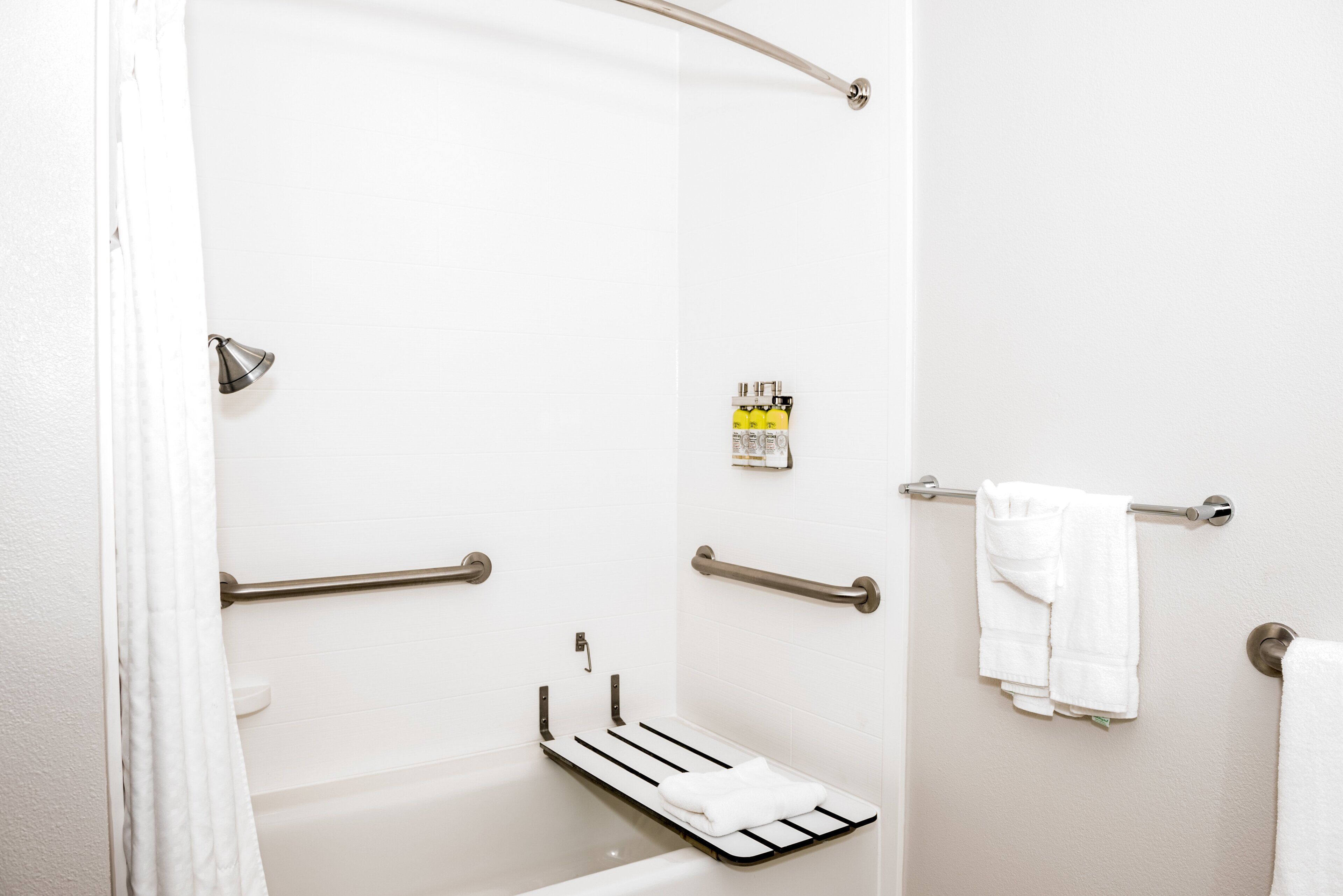 Une salle de bain équipée d’une douche, de barres d’appui, d’un porte-serviettes et d’une étagère contenant des articles de toilette.