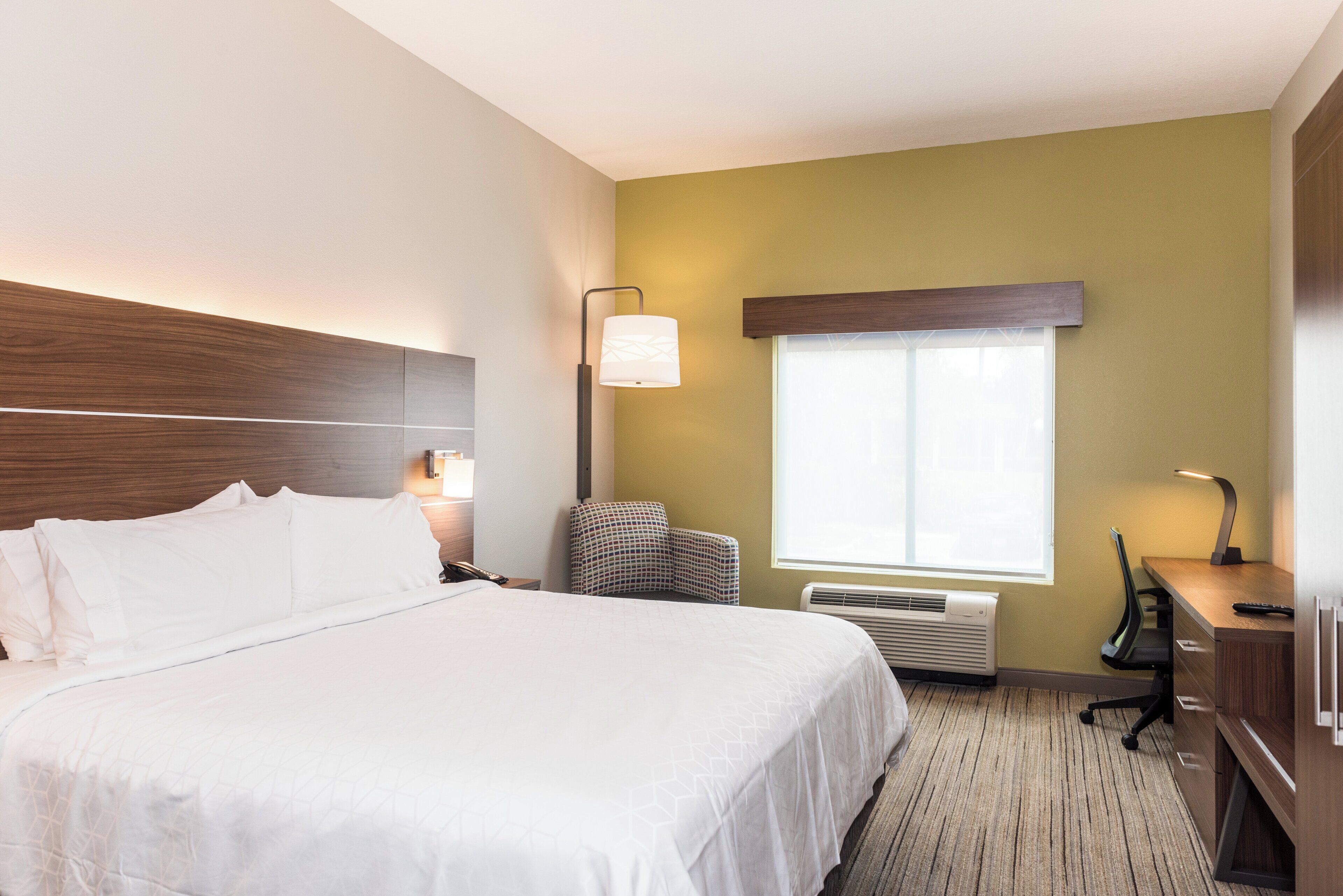 A hotel room with a large bed, bedside lamps, a desk with a chair, a window with blinds, and an air conditioning unit.