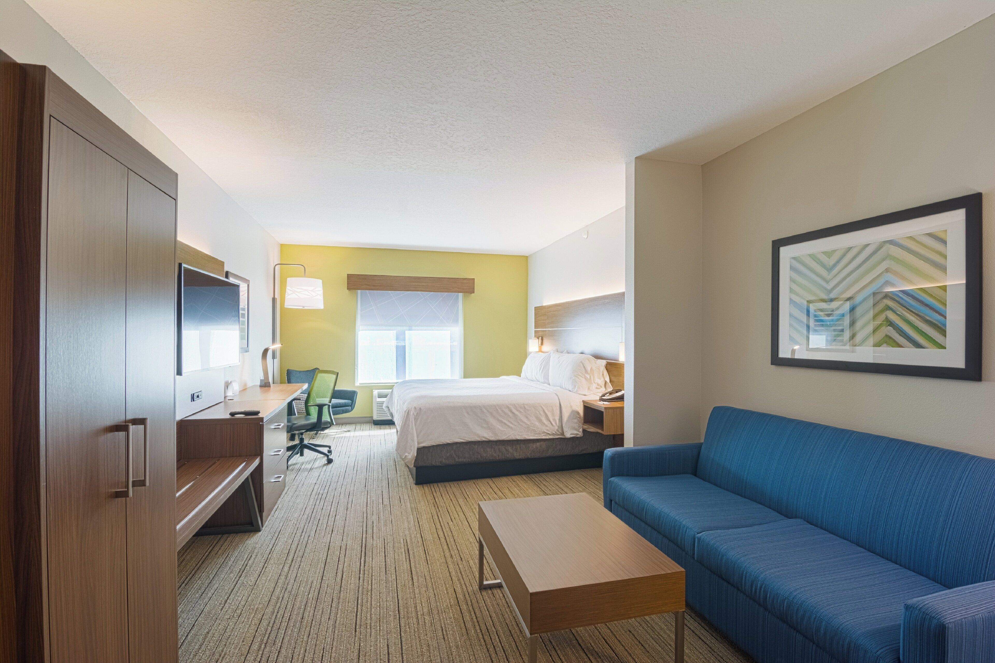 A hotel room with a bed, a blue sofa, a desk with a chair, a television, and a framed picture on the wall.