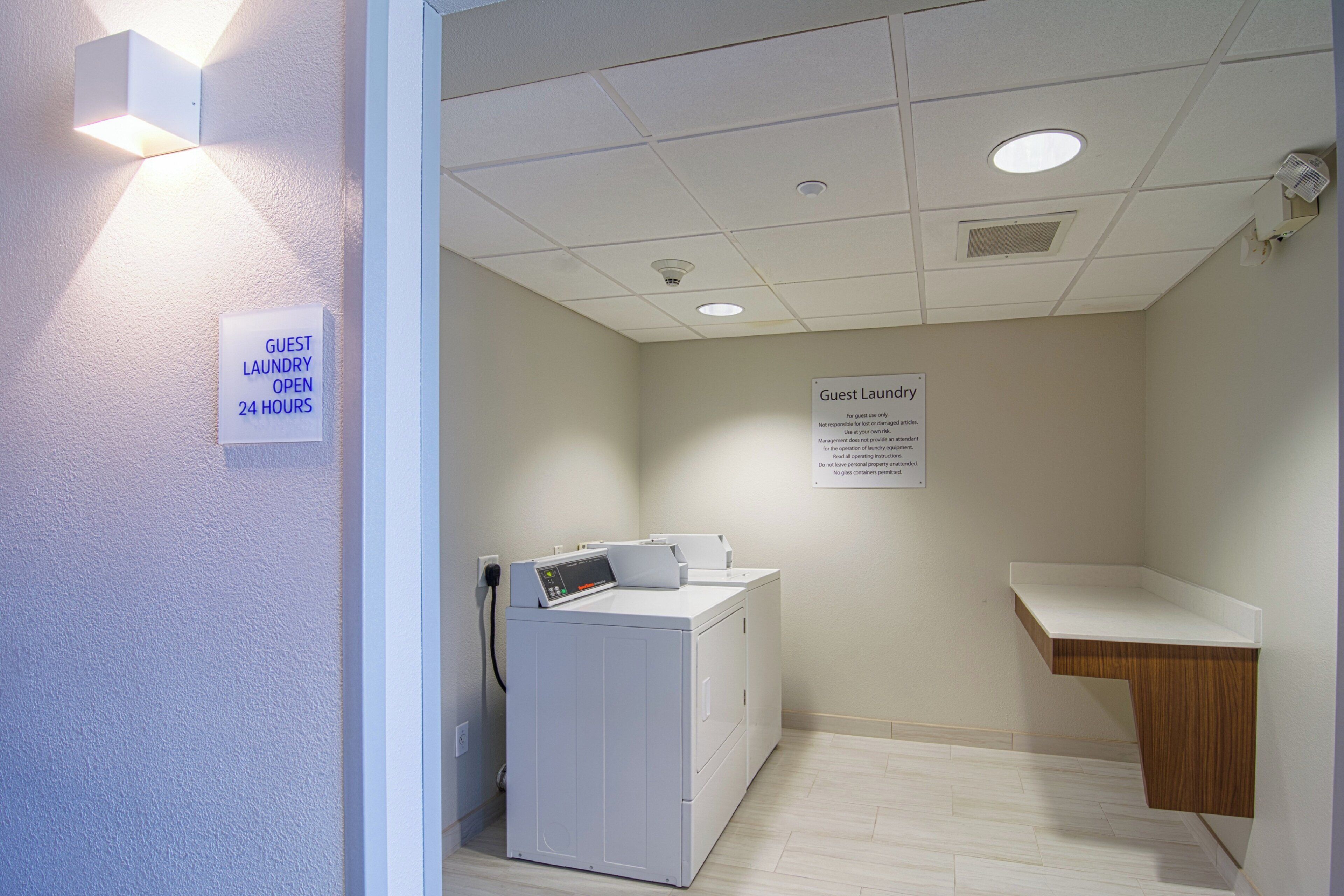 A laundry room with a washing machine, a sink, and signs indicating guest laundry is open 24 hours.