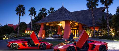 Two red sports cars parked in front of a resort with a thatched-roof structure.