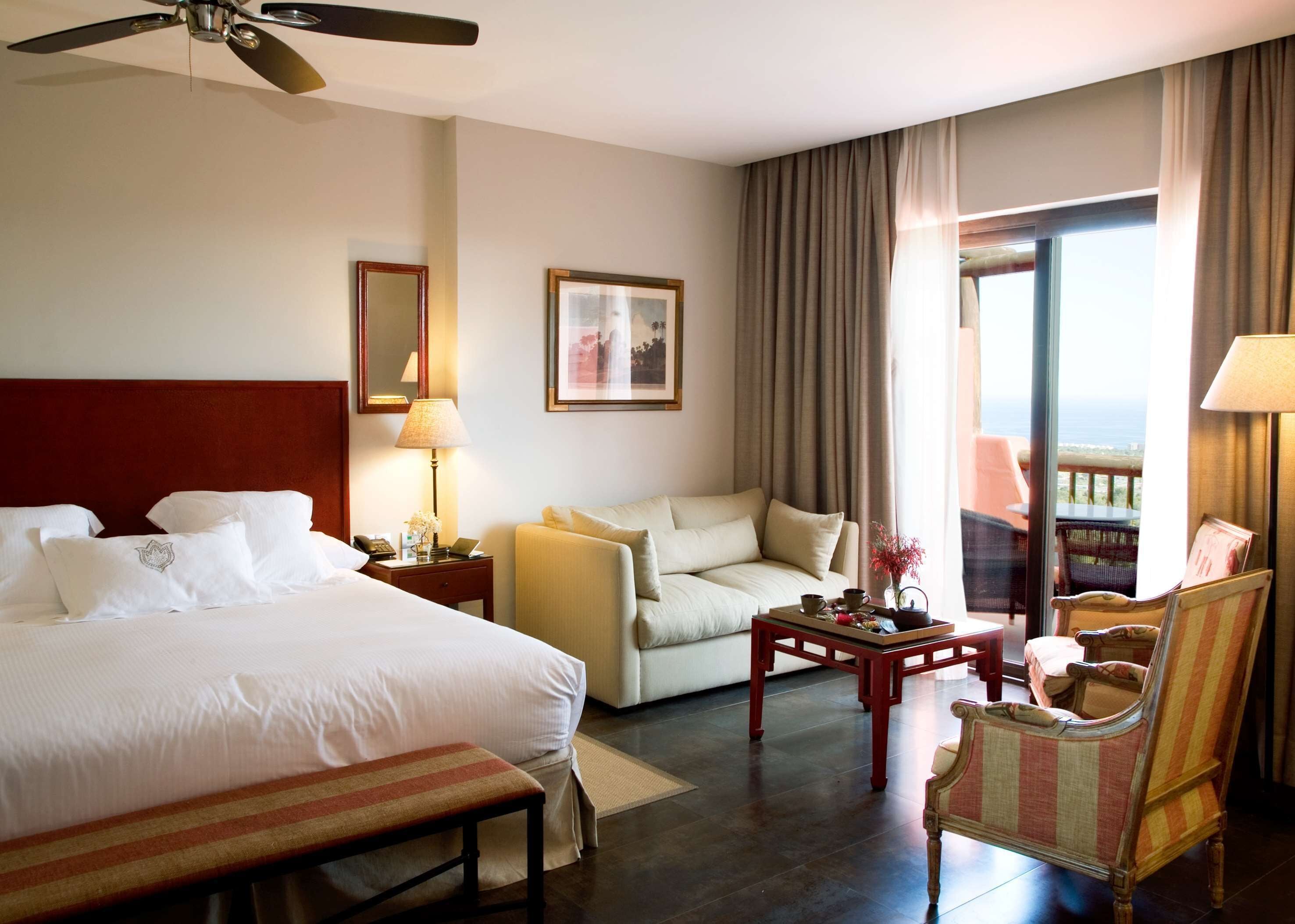 Habitación de hotel con cama, sofá, sillón, una mesita y balcón con vistas.