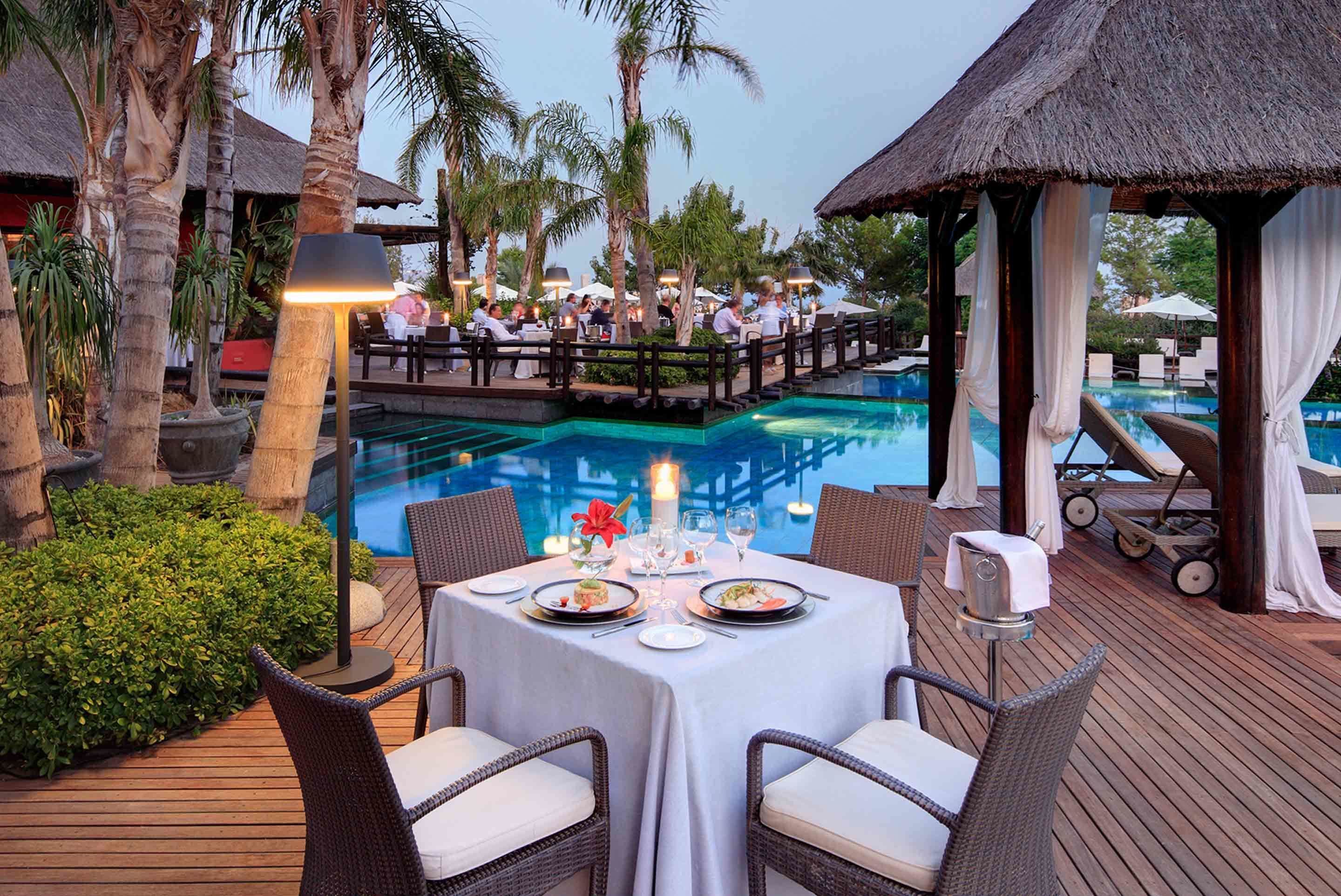 Un comedor con vista a la piscina, mesas dispuestas para dos personas y una estructura con techo de paja.