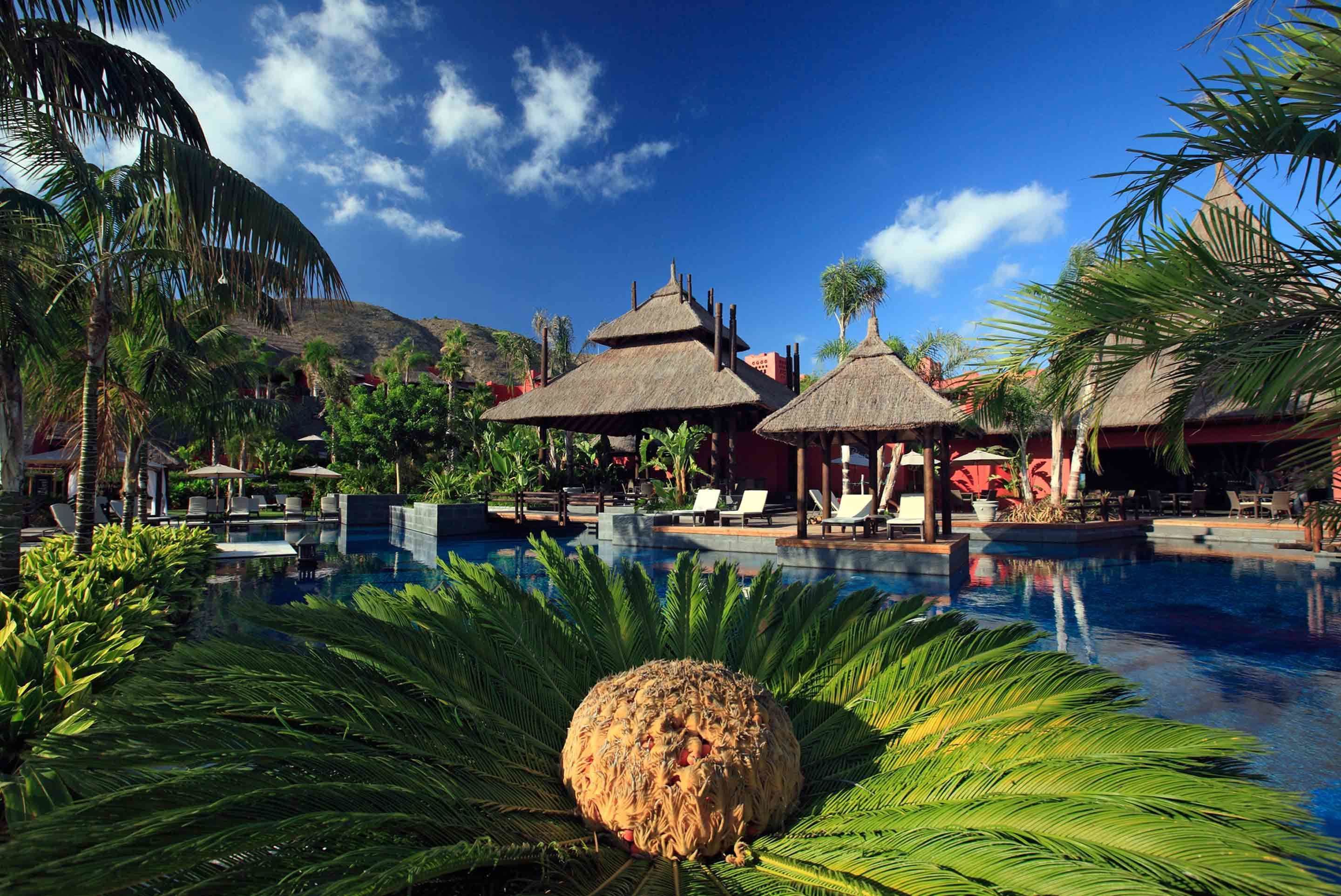 A pool area with lounge chairs, thatched-roof structures, and tropical plants.