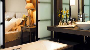 A hotel room with a bed, bedside table with a lamp, a large mirror, a bathroom with a sink and bathtub.