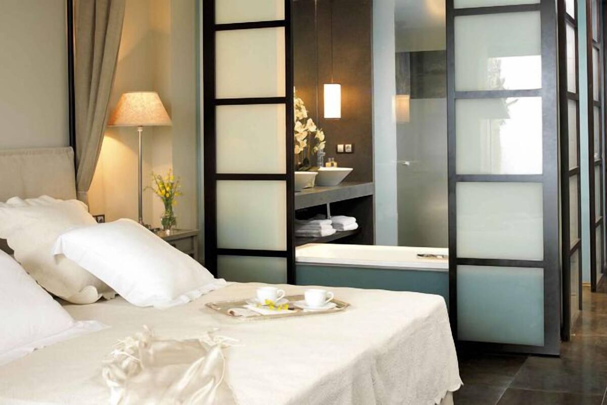 A hotel room with a bed, a bedside table with a lamp and flowers, a tray with cups and a flower, and a view of a bathroom with a sink and shelves.