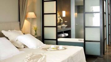 Una habitación de hotel con cama, mesita de noche con lámpara y flores, bandeja con tazas y una flor, y vistas a un baño con lavabo y estanterías.