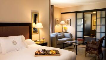 Habitación de hotel con una cama grande, un sofá, una mesa de centro y una silla. Hay una bandeja con alimentos para el desayuno sobre la cama.