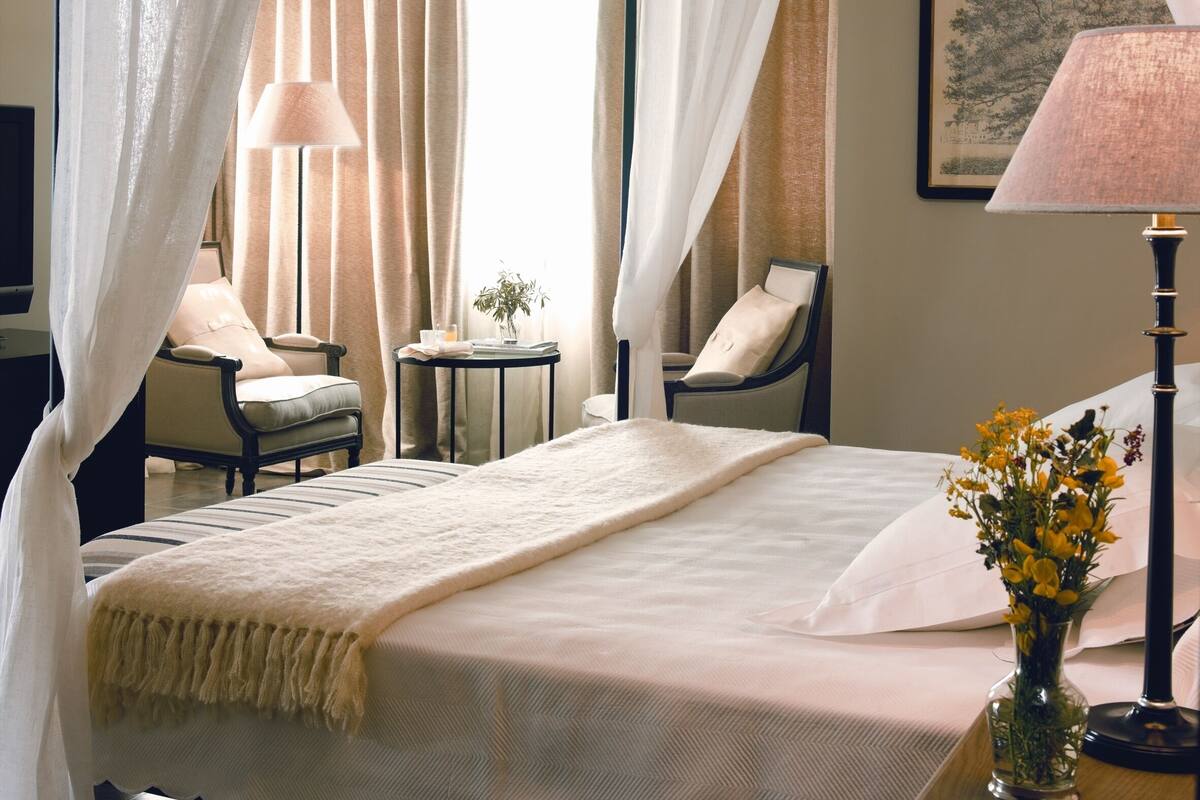 A hotel room with a bed, two chairs, a small table with a lamp, and a vase with flowers.