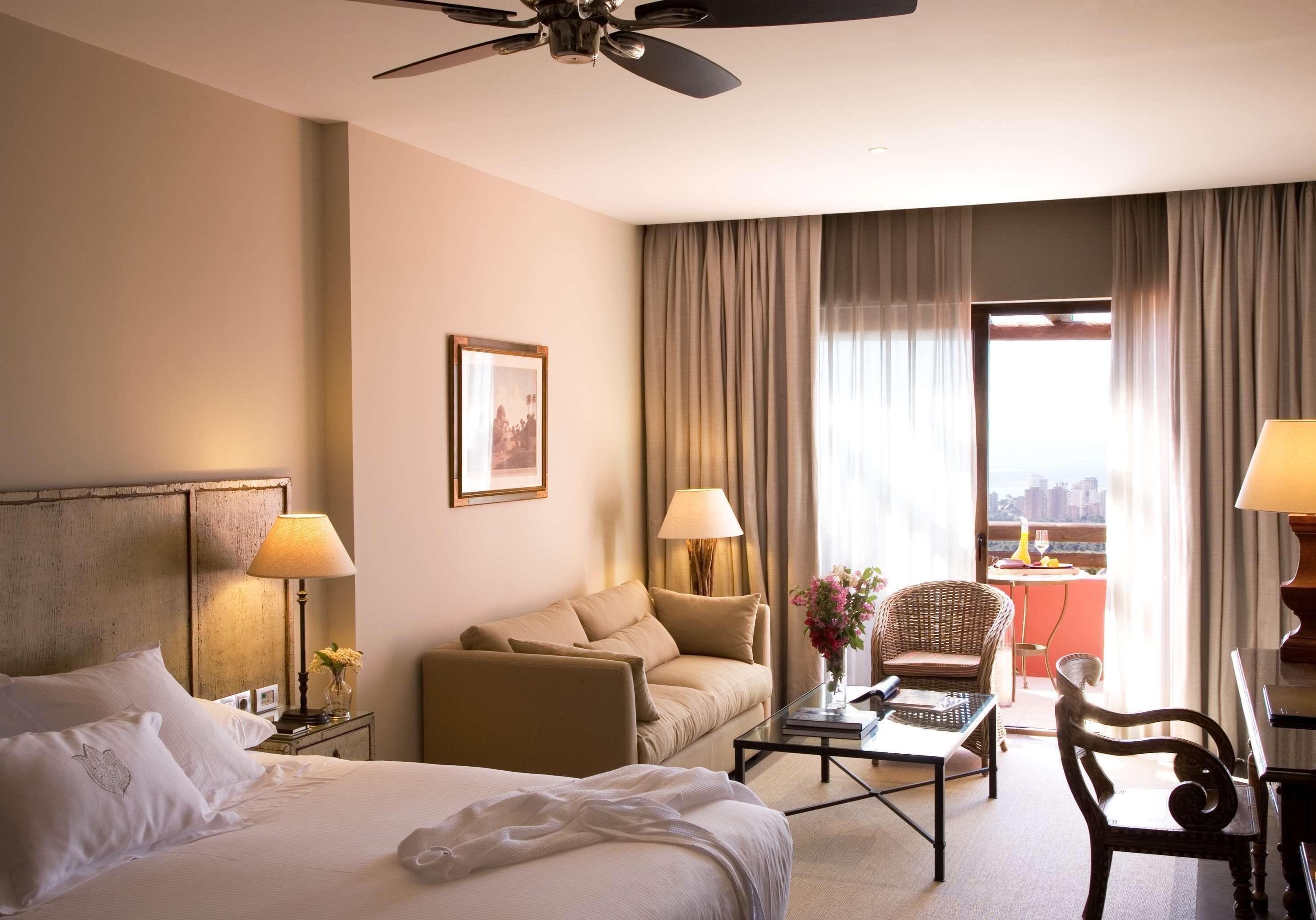 Habitación de hotel con una cama, un sofá, una silla, una mesita de centro y vistas a la ciudad a través de amplios ventanales.