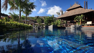 Una piscina cristalina con reflejos, rodeada de palmeras y estructuras con techo de paja.