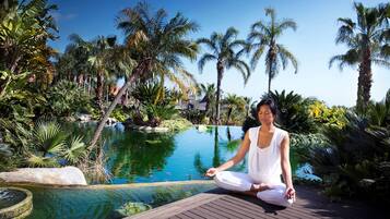 A person meditating by a pool surrounded by palm trees and tropical plants.