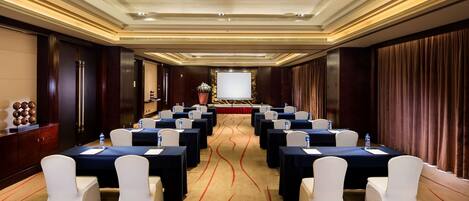 A conference room with rows of tables and chairs, a podium, and a large screen at the front.