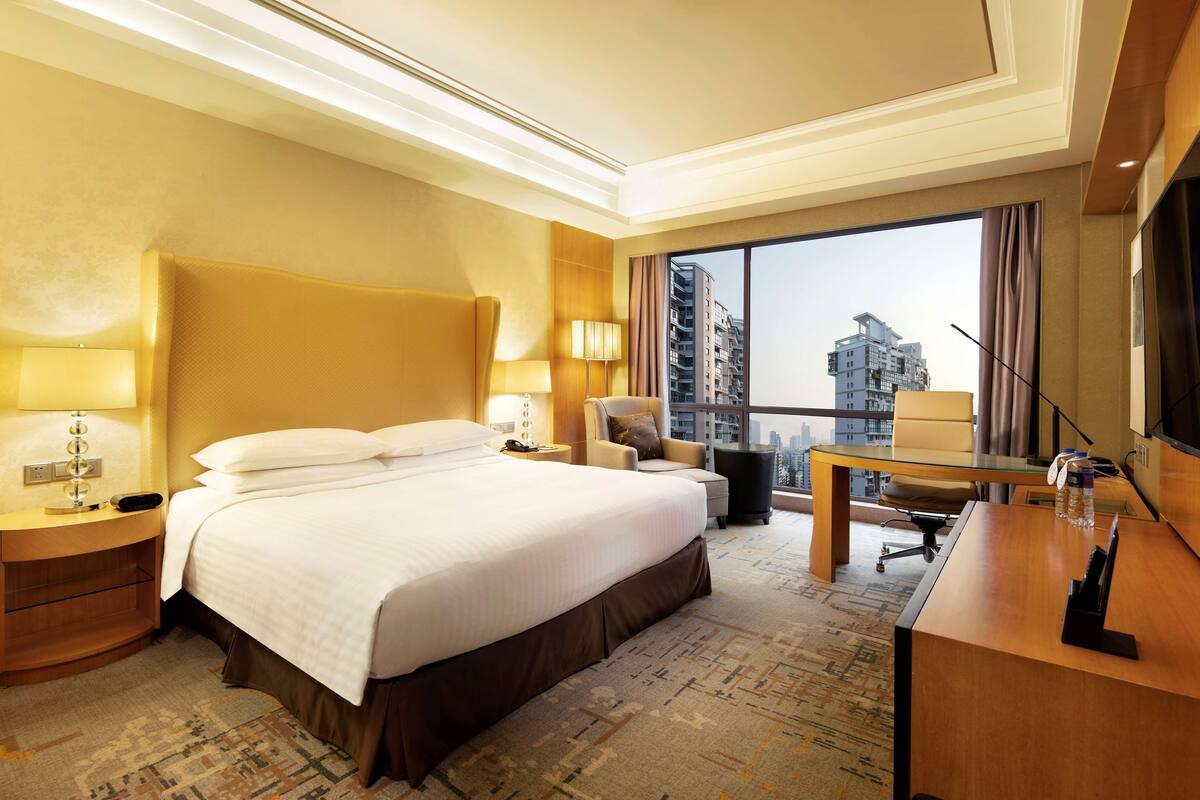 A hotel room with a large bed, a desk, a chair, a TV, and a view of the cityscape through the window.