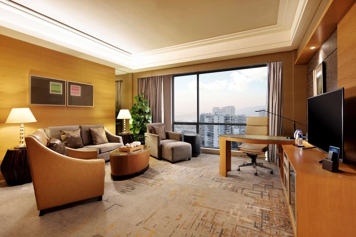 A modern hotel room with a sofa, armchair, desk, and large windows offering a city view.