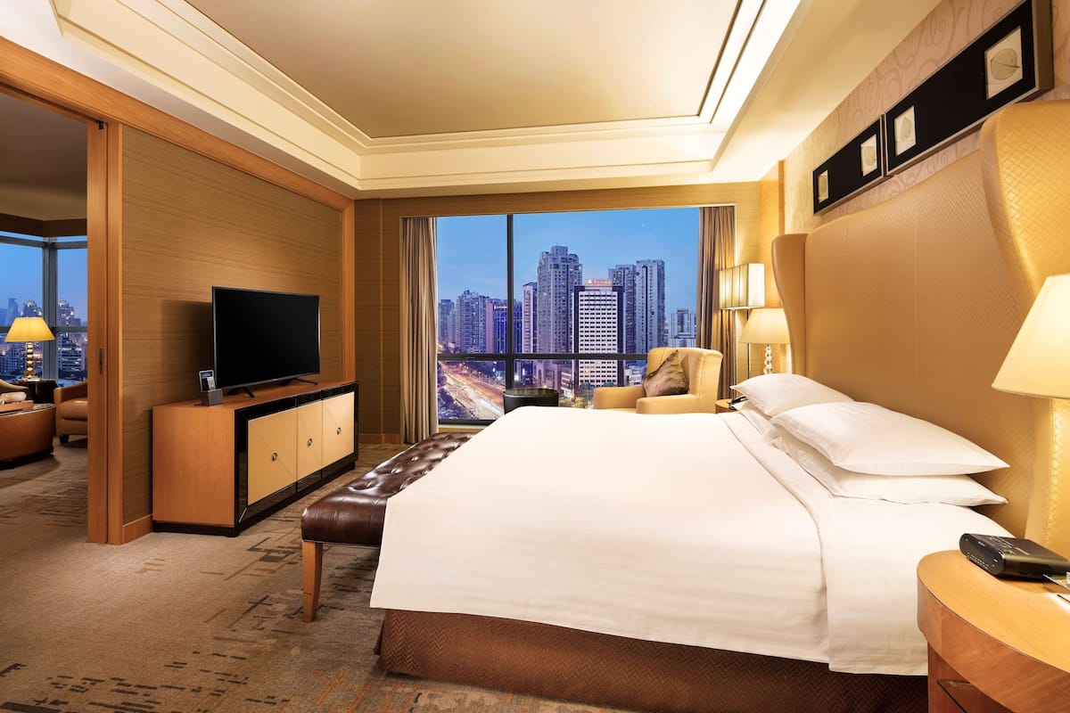 A hotel room with a large bed, a television on a wooden cabinet, a chair, a nightstand, and a view of the cityscape through large windows.