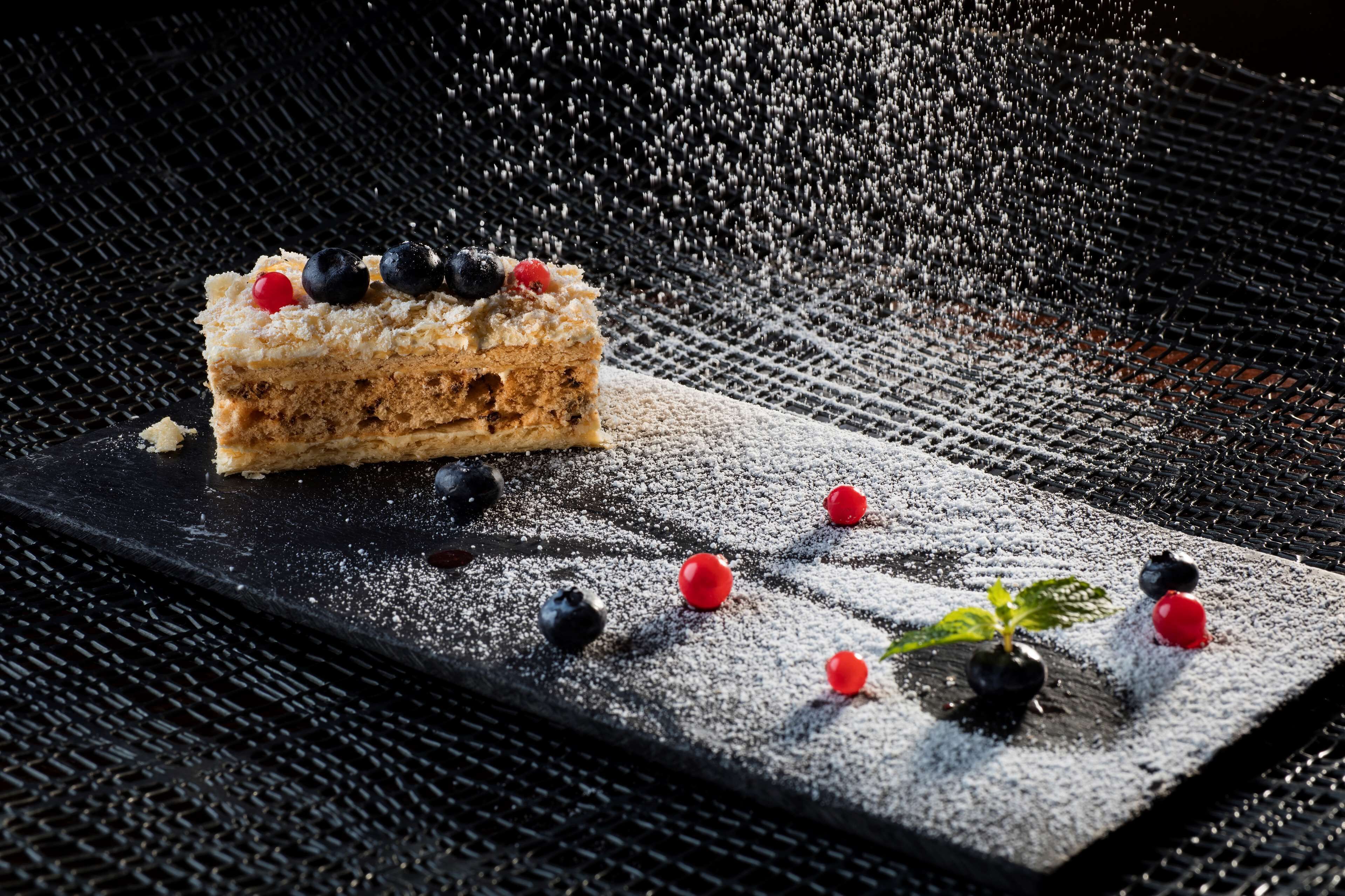 Une part de gâteau garnie de fruits rouges, présentée sur une assiette noire et saupoudrée de sucre glace, le tout posé sur une surface texturée.