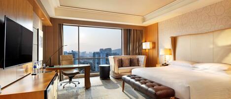 A hotel room with a large bed, a desk with a TV, a chair, a small table, and a view of the city.