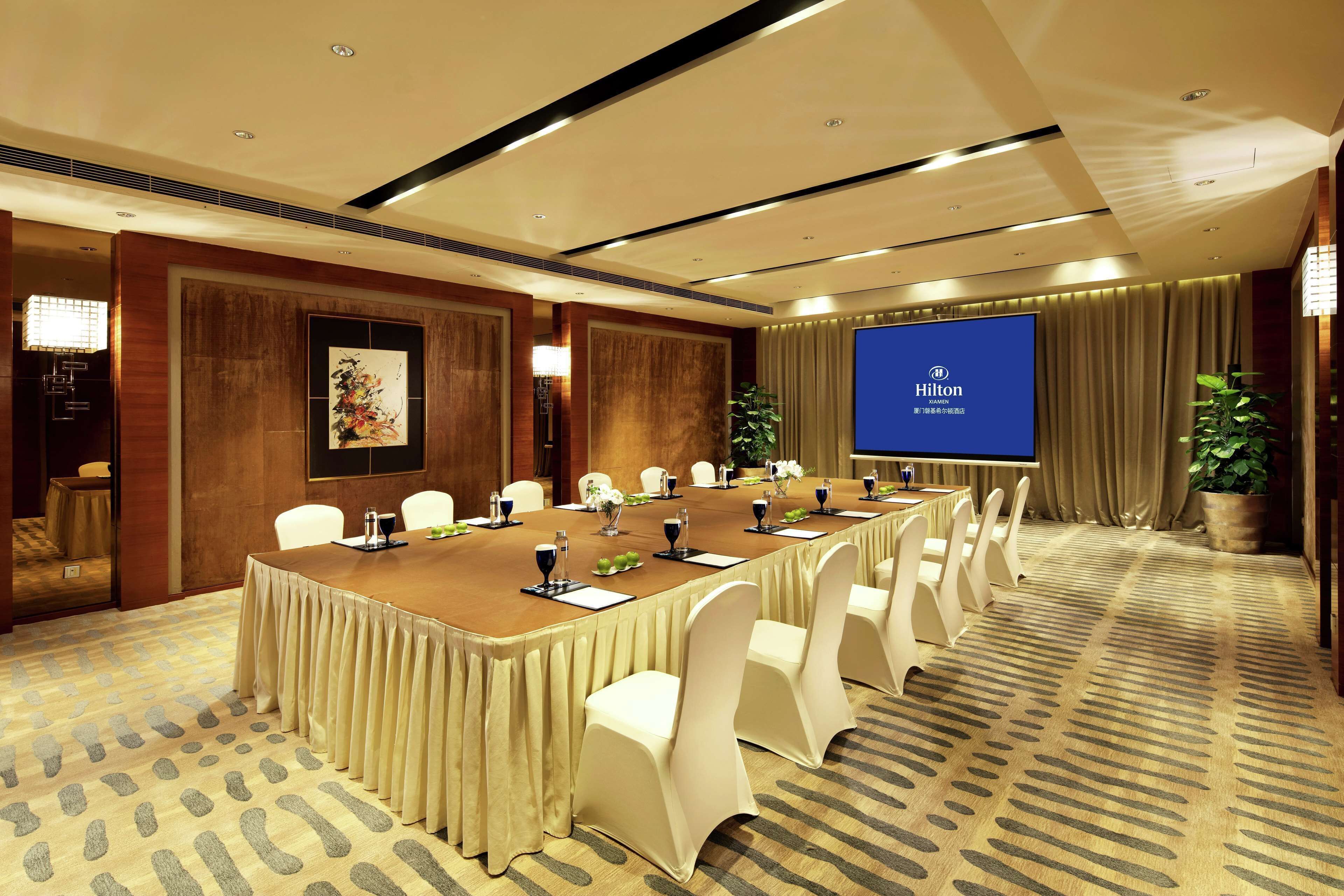 A conference room with a long table, white chairs, a projector screen displaying 'Hilton', and a patterned carpet.