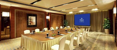 Un salón de conferencias con una larga mesa, sillas blancas, una pantalla de proyector mostrando 'Hilton' y una alfombra estampada.
