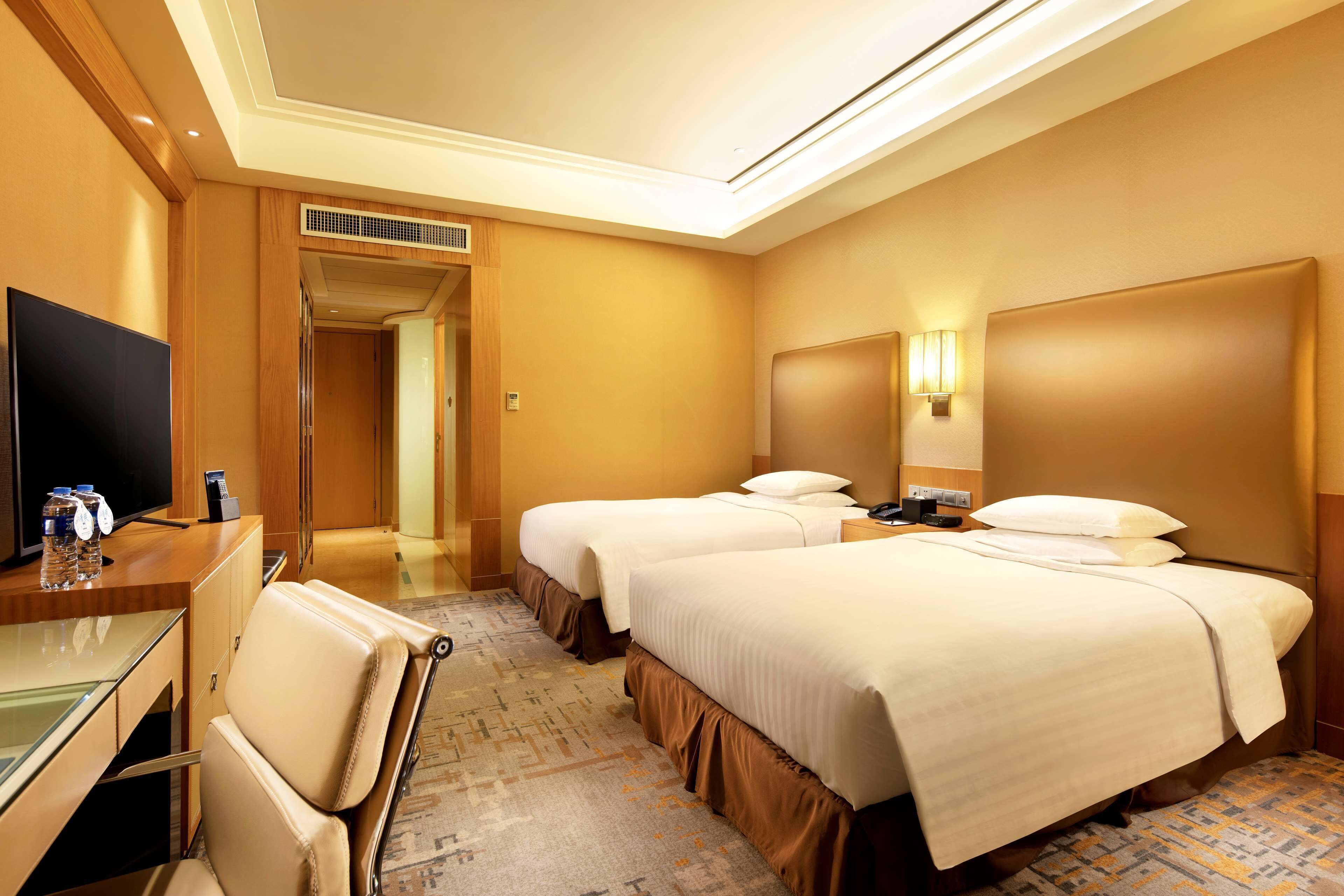 A hotel room with two beds, a desk with a flat-screen TV, a chair, and a carpeted floor.