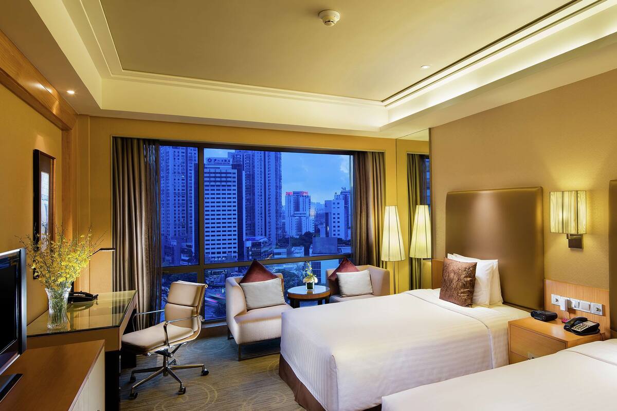 A hotel room with two beds, a desk, a chair, a TV, and a view of the cityscape.