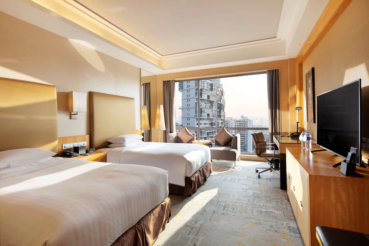 A hotel room with two beds, a desk, a TV, and a view of a cityscape.