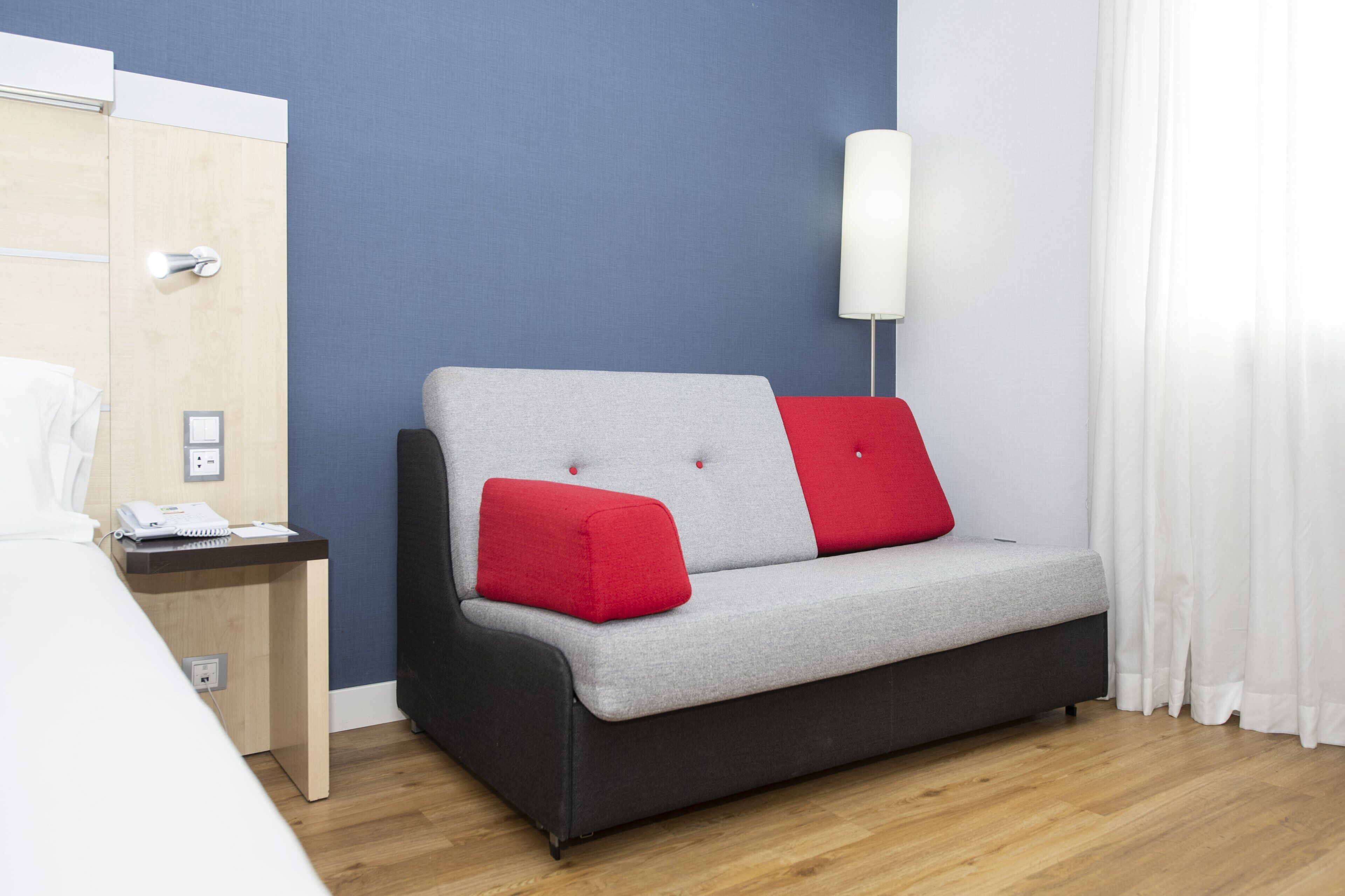 A modern hotel room with a grey sofa, red cushions, a wooden nightstand, a white lamp, and a bed with white linens.