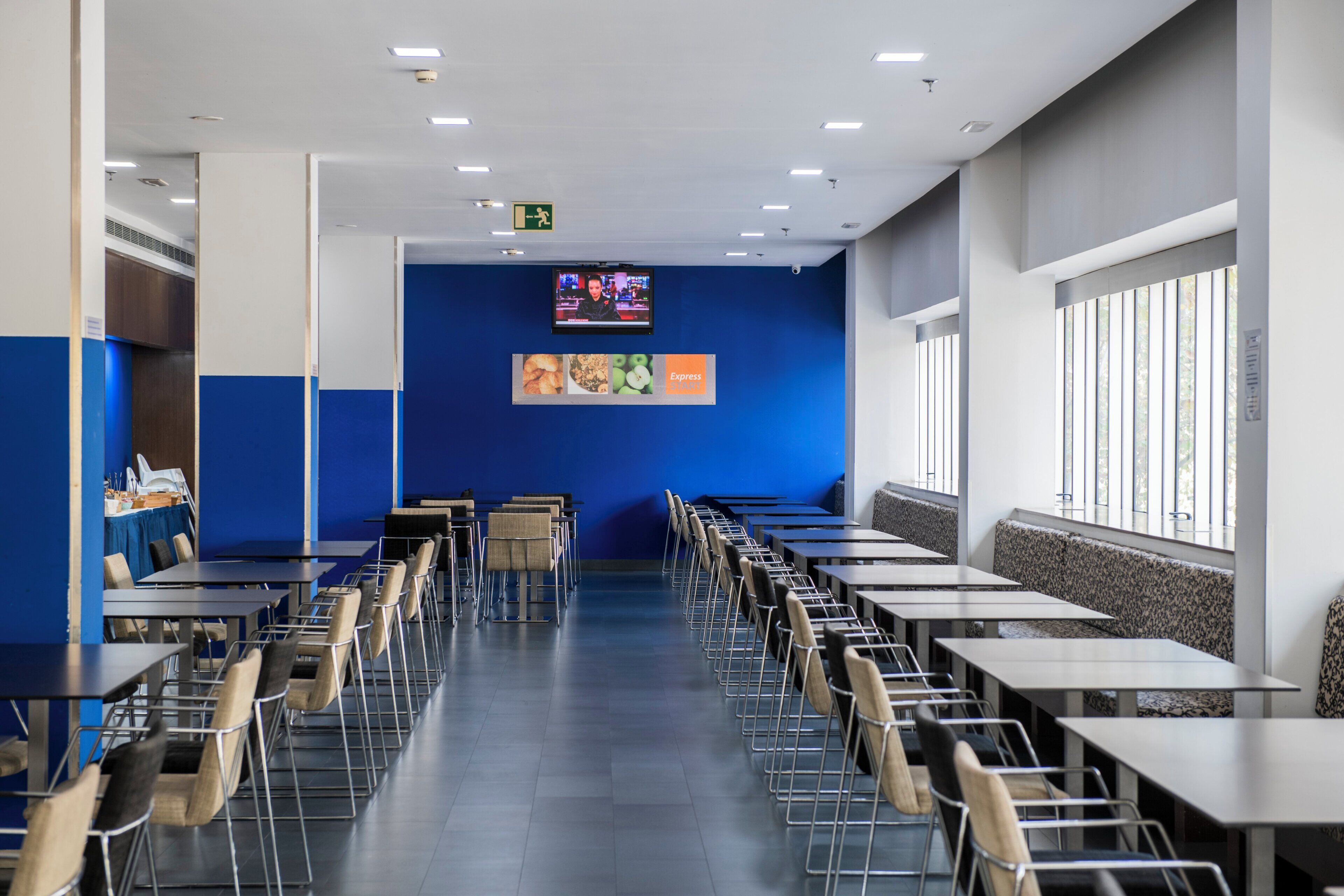 A cafeteria with long tables and chairs, a television screen on the wall, and a window on the right side.