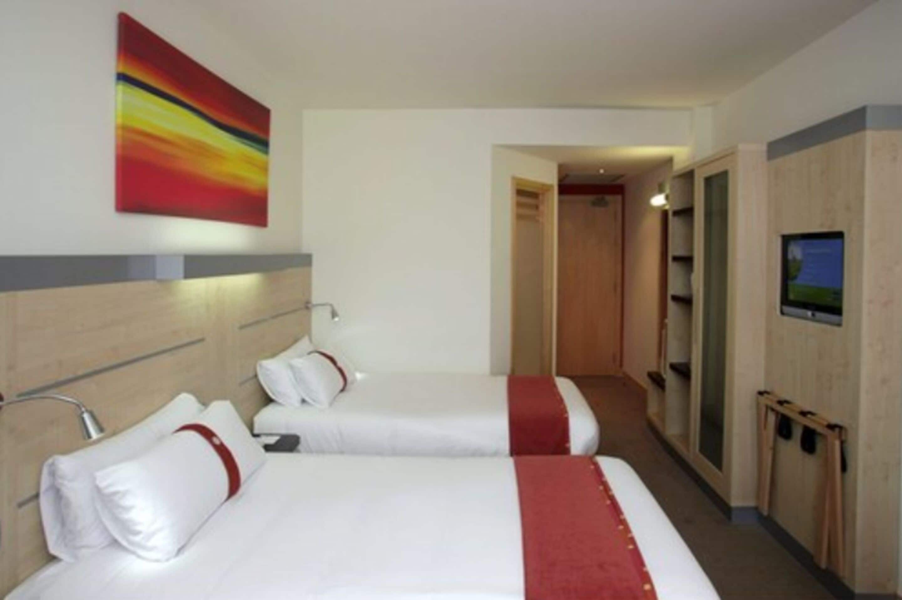 A hotel room with two beds, a red and white striped painting, a television, and a wooden wardrobe.