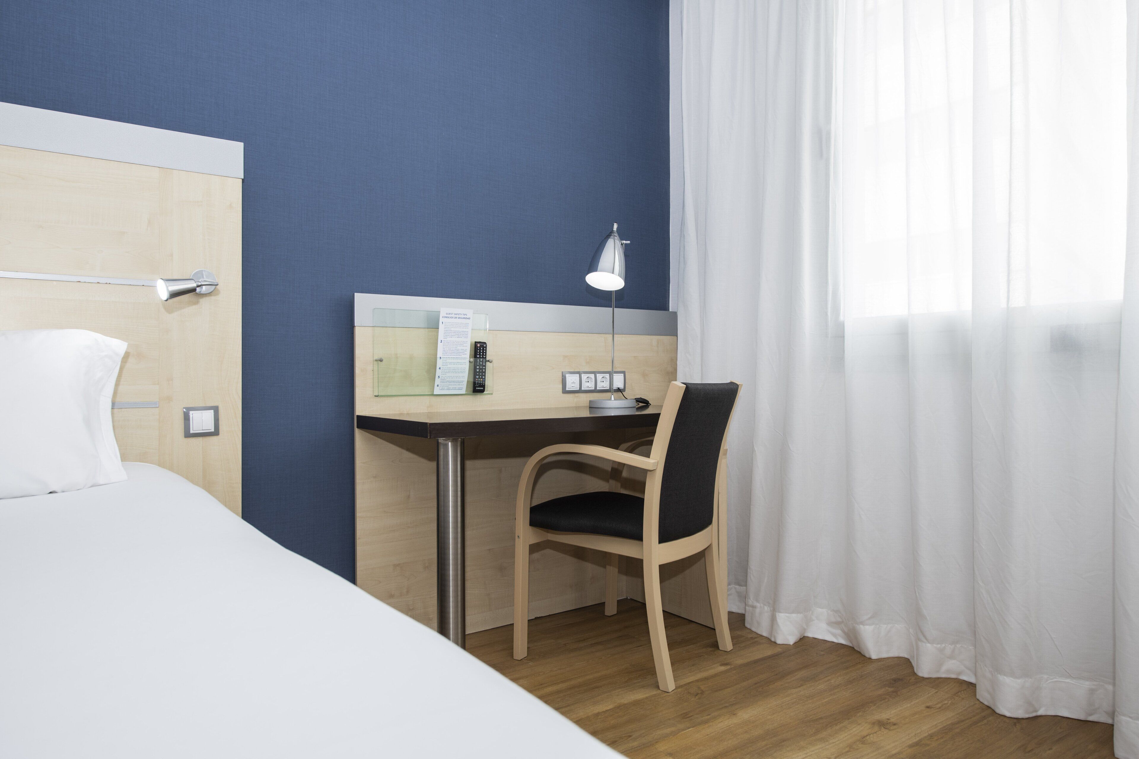 Une chambre d’hôtel avec un lit, un bureau, une chaise, une lampe et une fenêtre avec des rideaux.