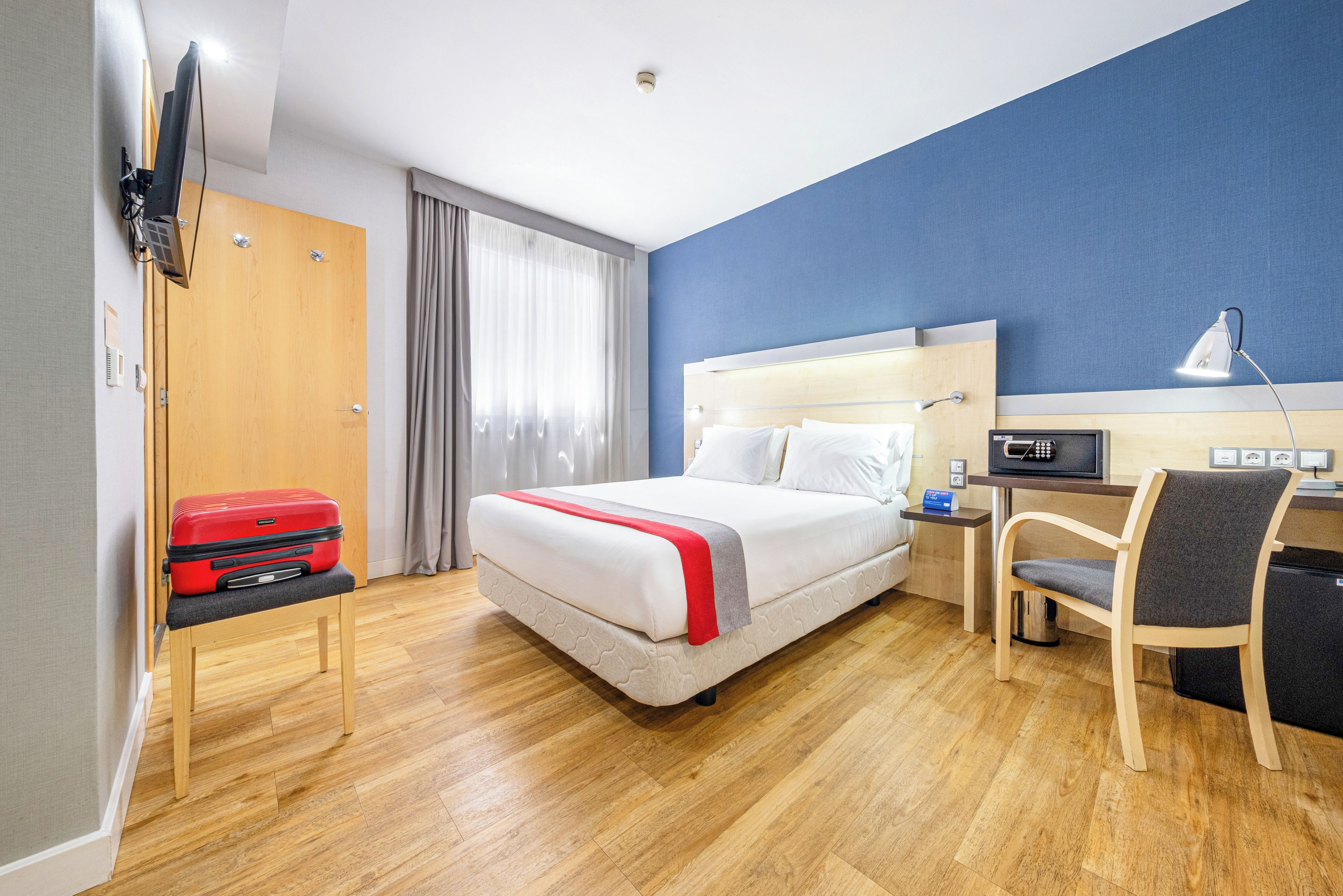 A hotel room with a bed, a desk, a chair, a TV, and a suitcase.