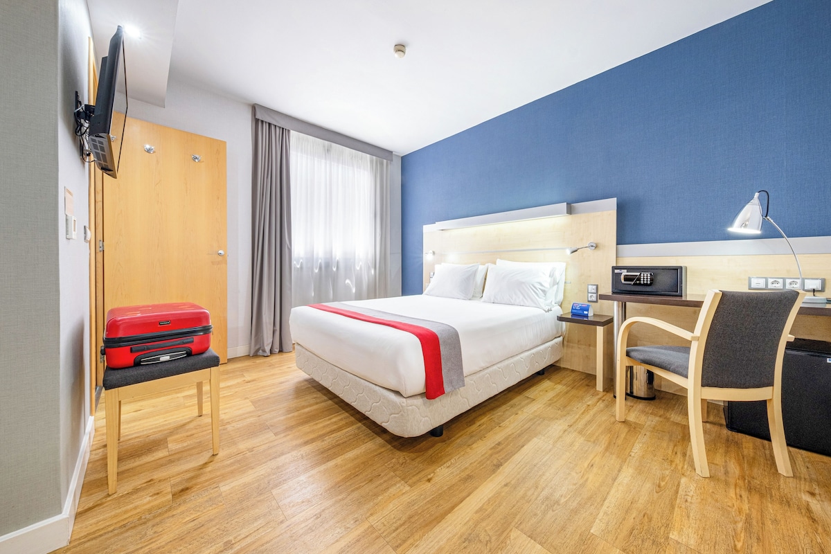 A hotel room with a bed, a desk, a chair, a TV, and a suitcase.