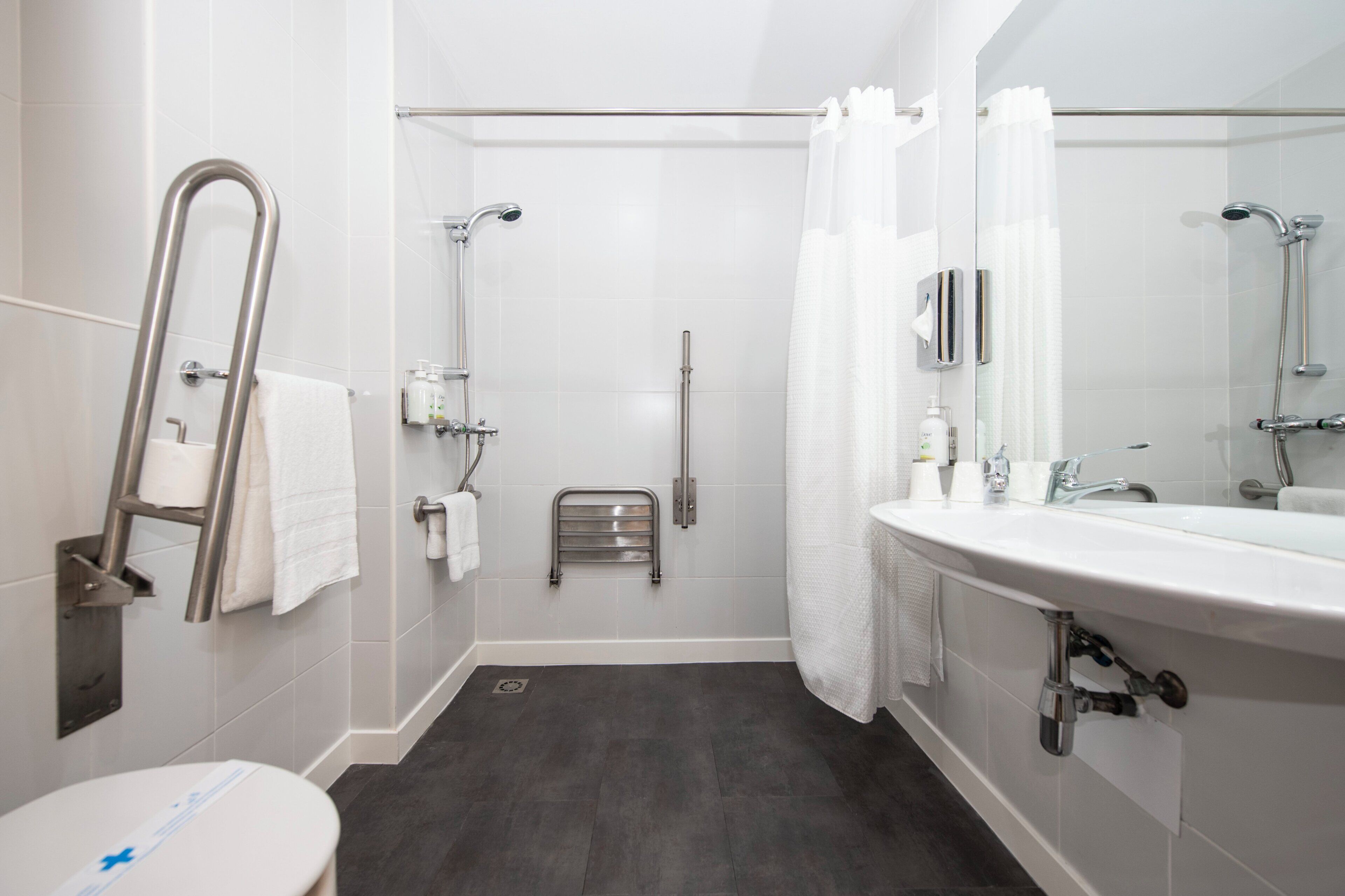 Un baño moderno con lavamanos, espejo y ducha blancos. Incluye una barra de apoyo, un perchero y un calentador pequeño para toallas.