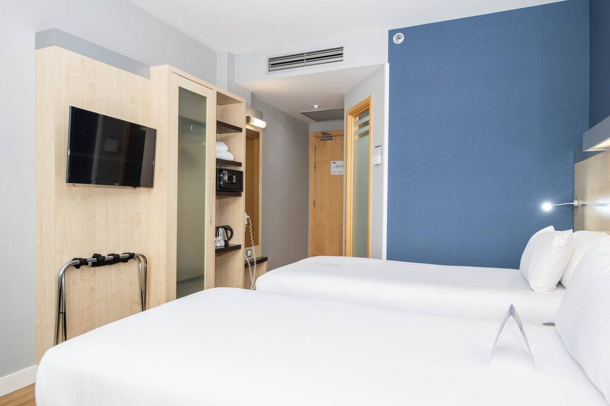 A hotel room with two beds, a TV mounted on the wall, a microwave, and a wooden wardrobe.