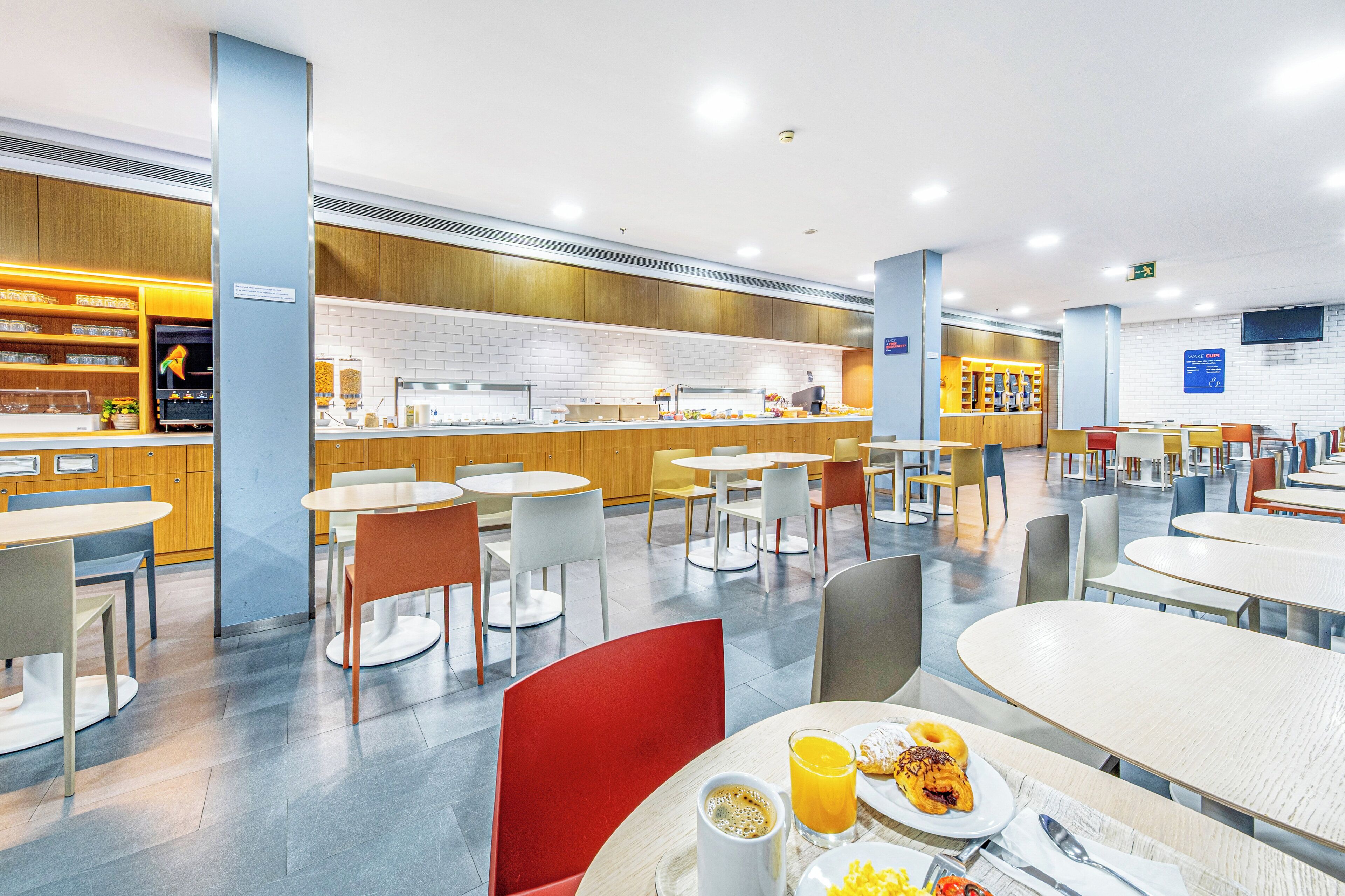 A modern dining area with tables and chairs, a counter with food items, and a kitchen in the background.