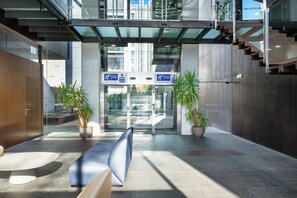 Eine moderne Hotelhalle mit Glastür, zwei Zimmerpflanzen, einer blauen Bank und einer Treppe.