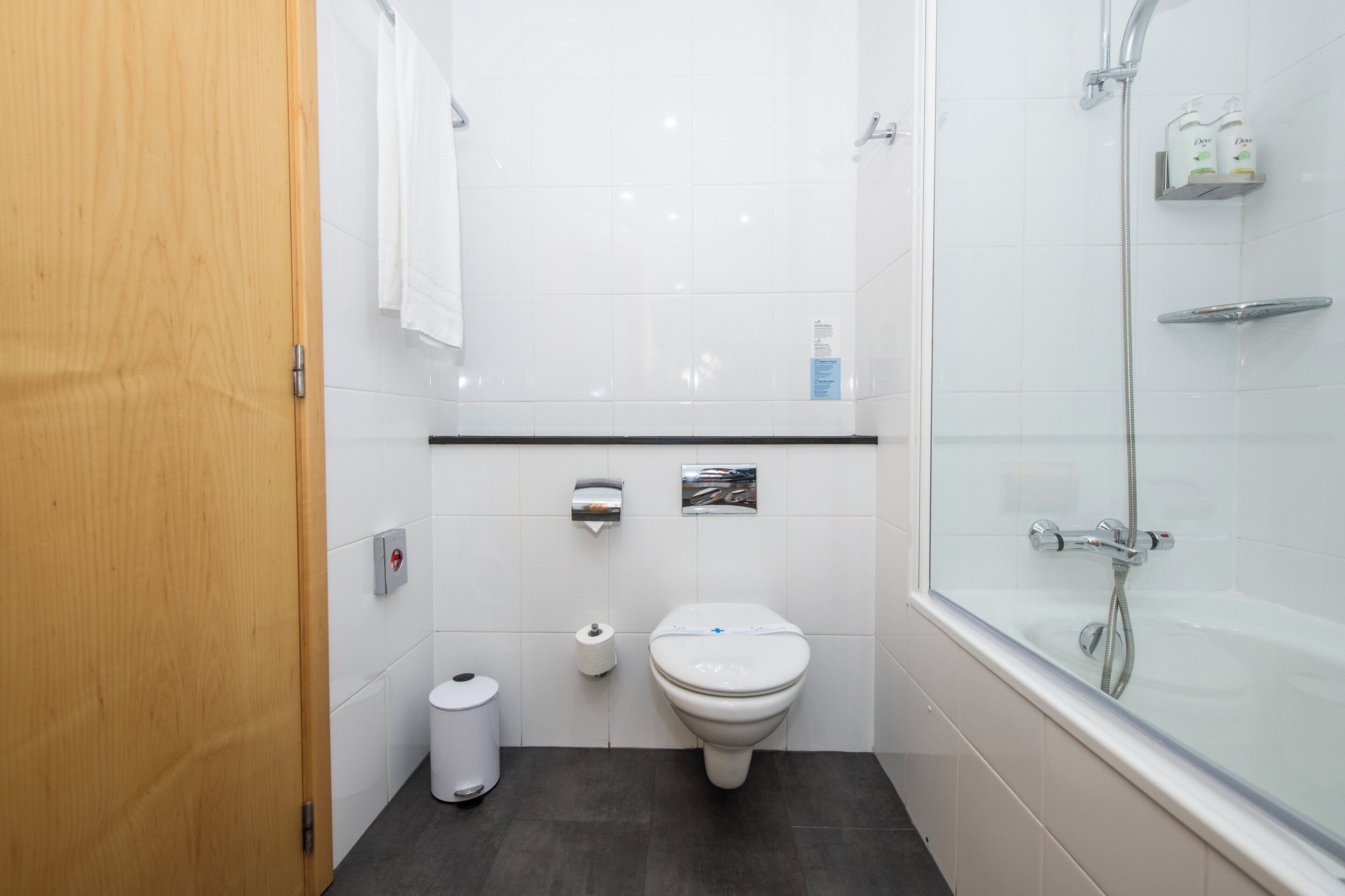 Une salle de bain moderne équipée de toilettes, d’un distributeur d’essuie-tout, d’une cabine de douche en verre et d’une porte en bois.