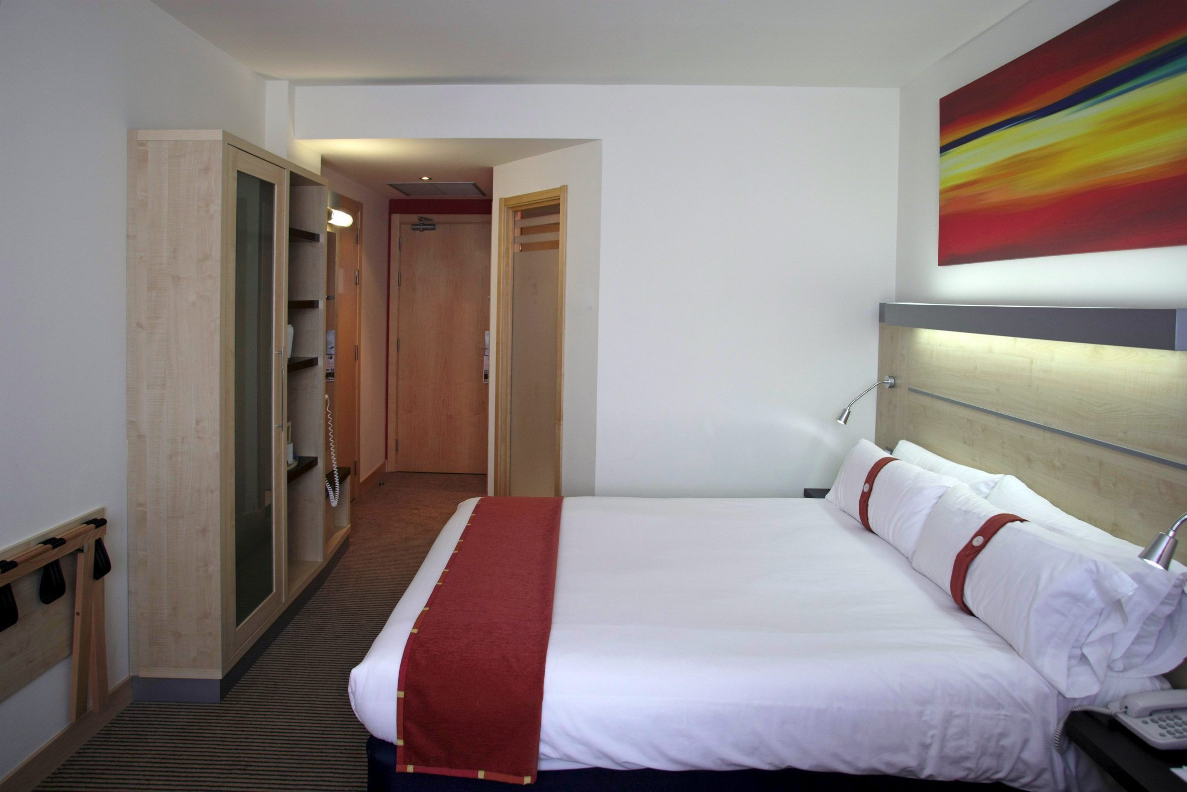 A hotel room with a large bed, a wooden wardrobe, a walk-in closet, and a modern abstract painting on the wall.