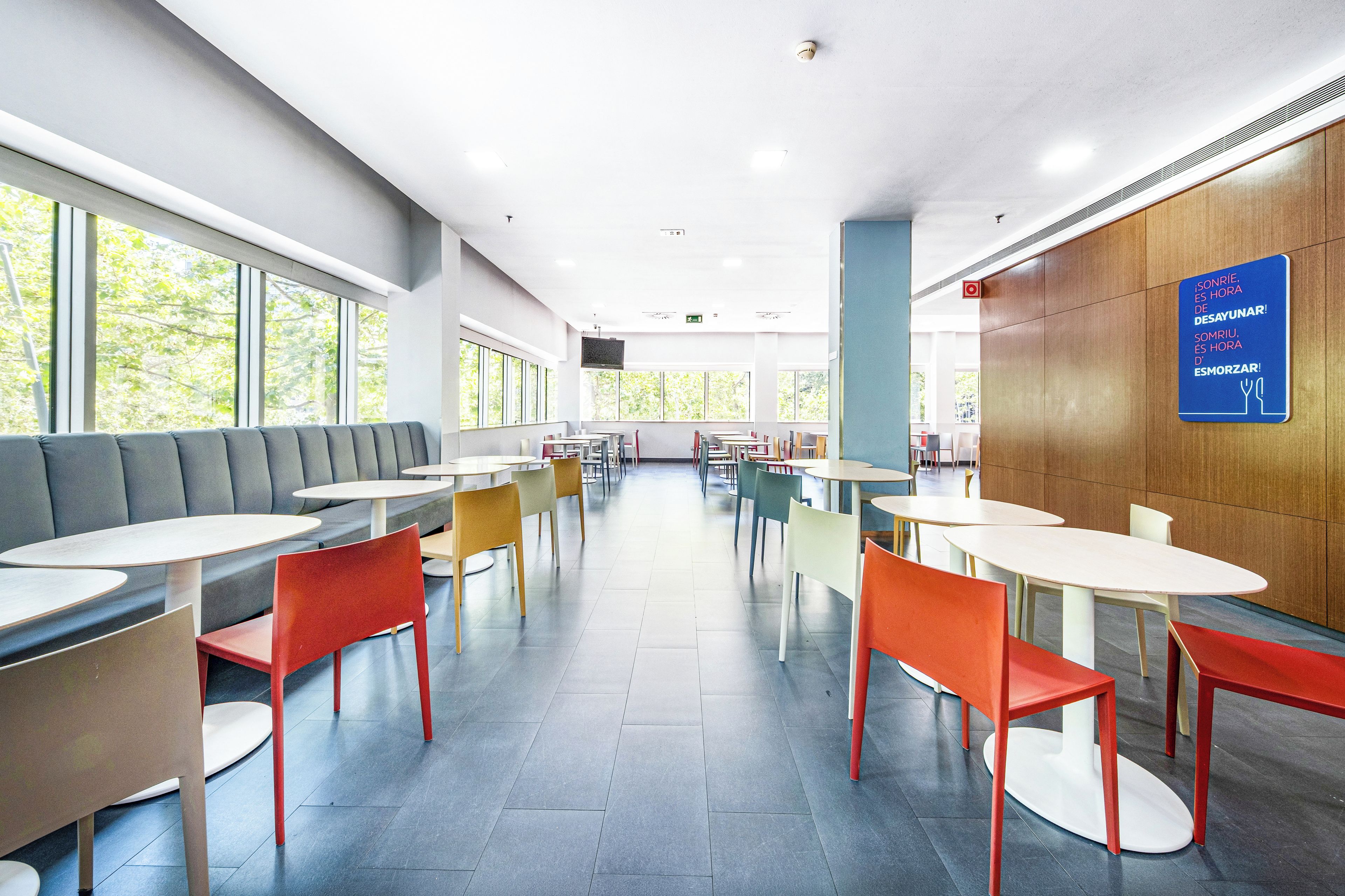 Une cafétéria moderne avec des tables et des chaises hautes, de grandes fenêtres et une enseigne bleue avec du texte.