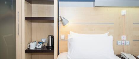 Una habitación de hotel con una cama, una mesita de noche con lámpara, un teléfono y un estante con hervidor y cafetera.