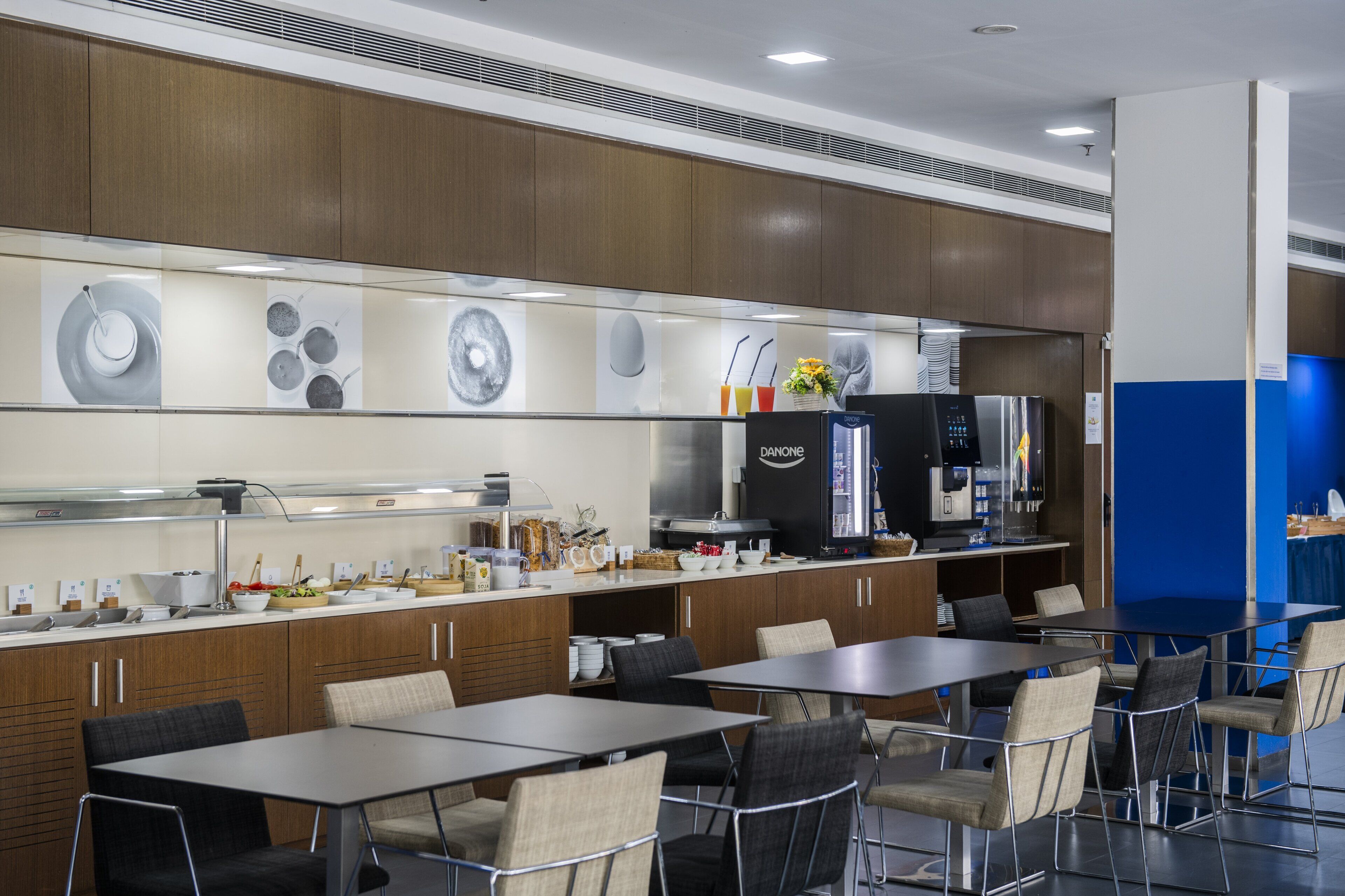 A modern kitchen with wooden cabinets, a stainless steel refrigerator, and a coffee machine. There are several tables and chairs arranged for dining.
