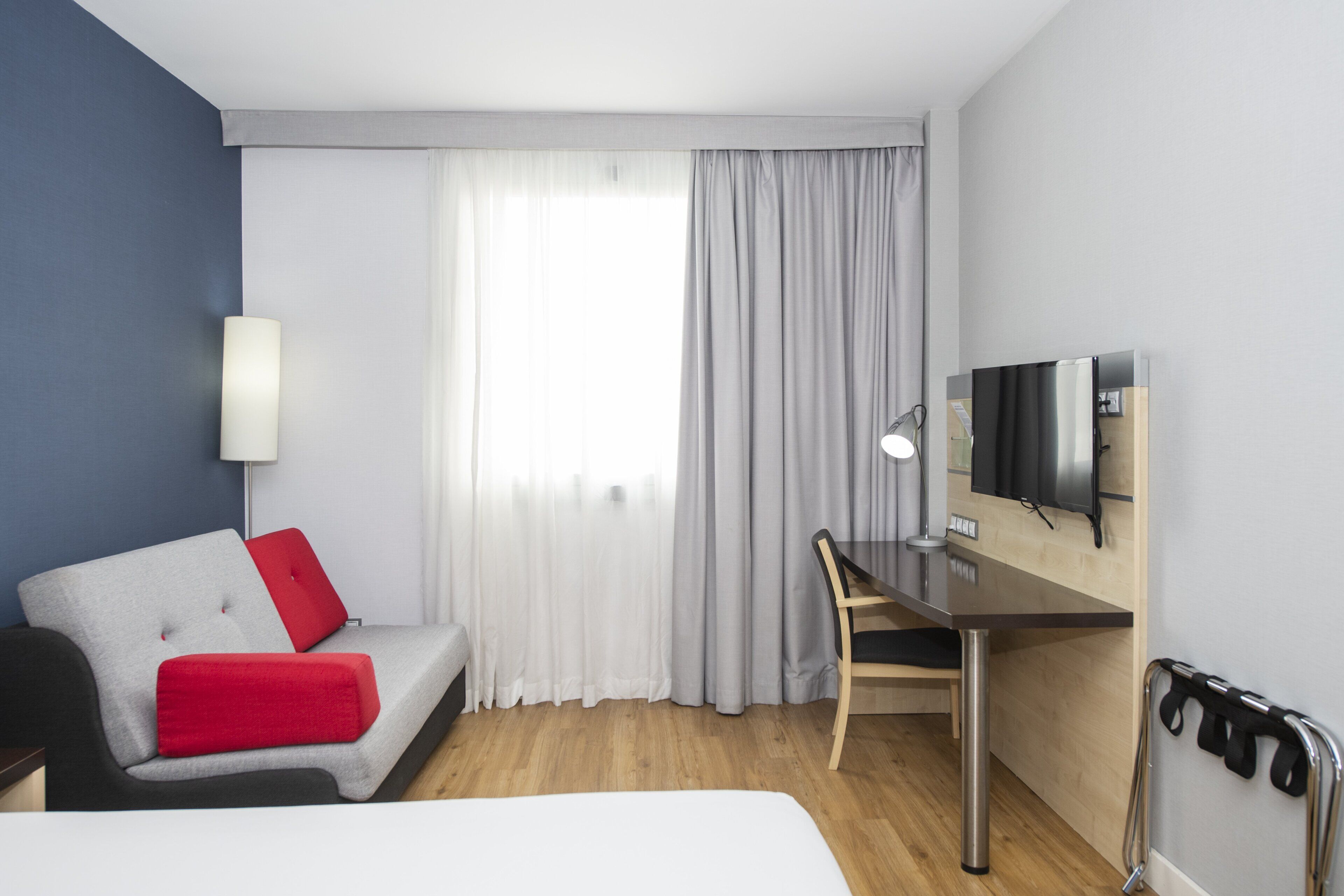 A hotel room with a bed, a desk, a chair, a TV, and a window with curtains.